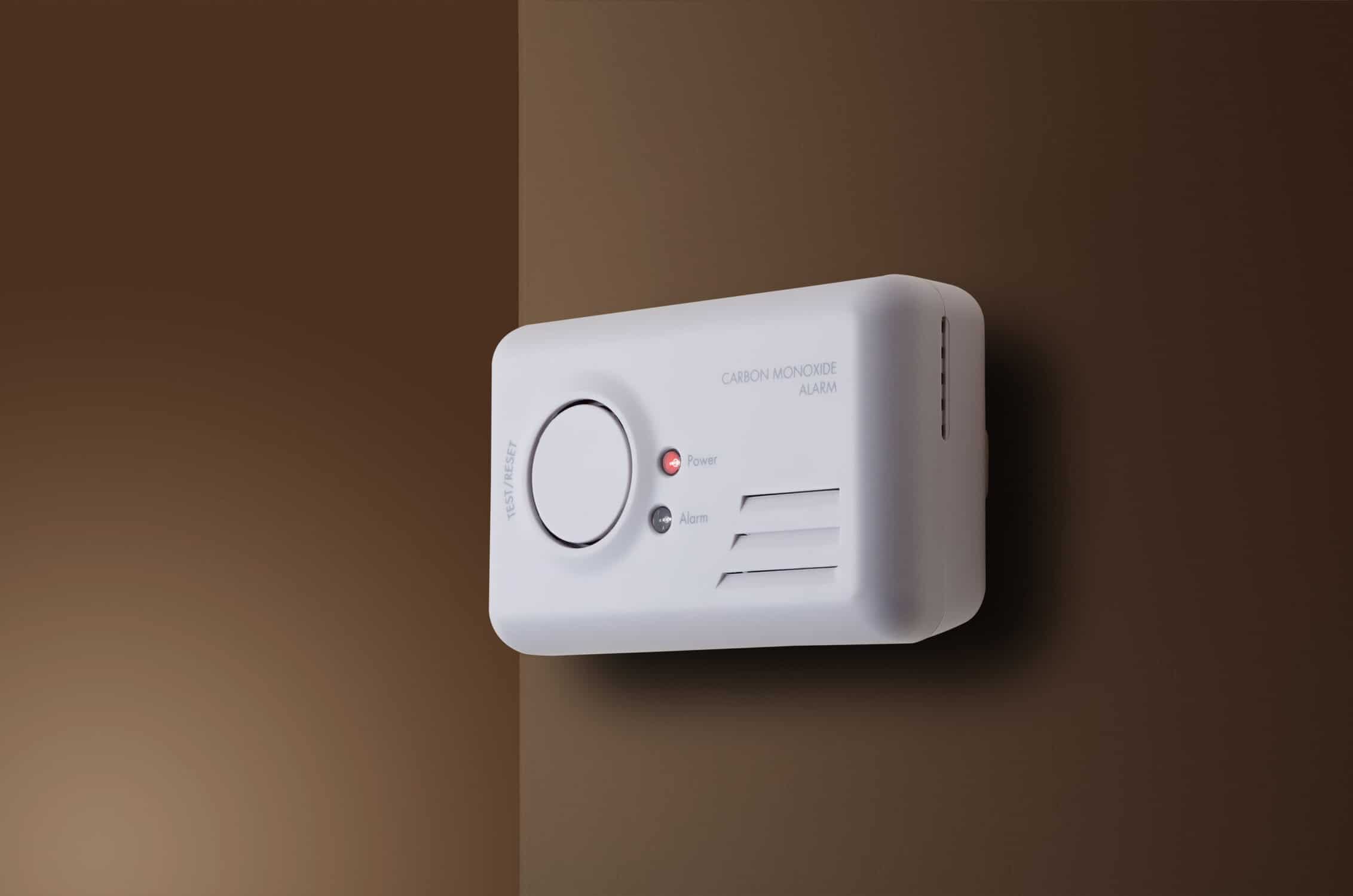 A White Carbon Monoxide Alarm Is Mounted On A Two-Tone Brown Wall, Showing Power And Alarm Indicator Lights.