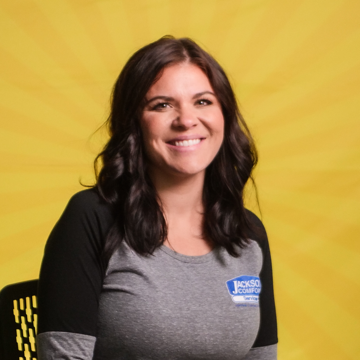 A Woman With Wavy Brown Hair Sits On A Chair, Smiling, Representing Our Team In A Gray And Black &Quot;Jackson Comfort&Quot; Shirt, Against A Yellow Background With A Radiating Pattern.