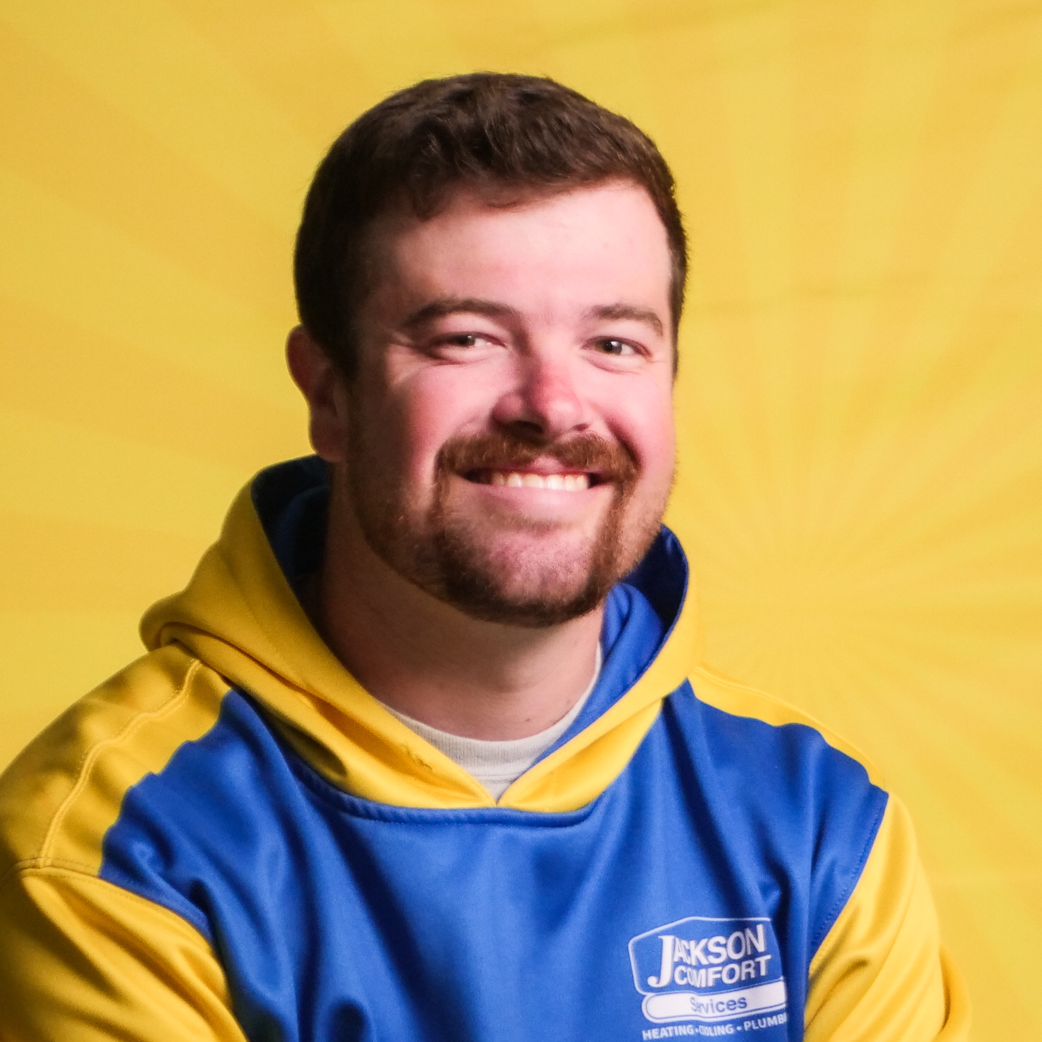 A Man With Short Brown Hair And A Beard Smiles At The Camera, Representing Our Team In A Blue And Yellow &Quot;Jackson Comfort Services&Quot; Hoodie, Set Against A Vibrant Yellow Background.