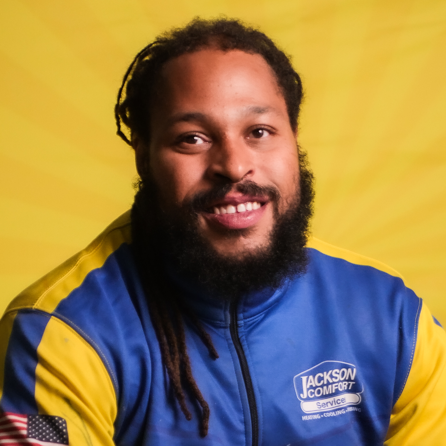 A Man With Long Hair And A Beard, Representing Our Team, Sits In Front Of A Yellow Background Wearing A Blue And Yellow Jackson Comfort Service Jacket.