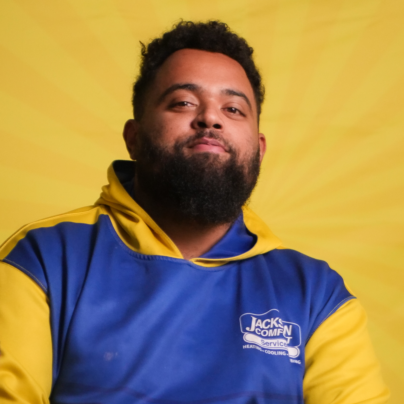 A Man With A Beard, Representing Our Team, Wears A Blue And Yellow &Quot;Jack'S Comfort Service&Quot; Hoodie As He Sits Against A Bright Yellow Background.
