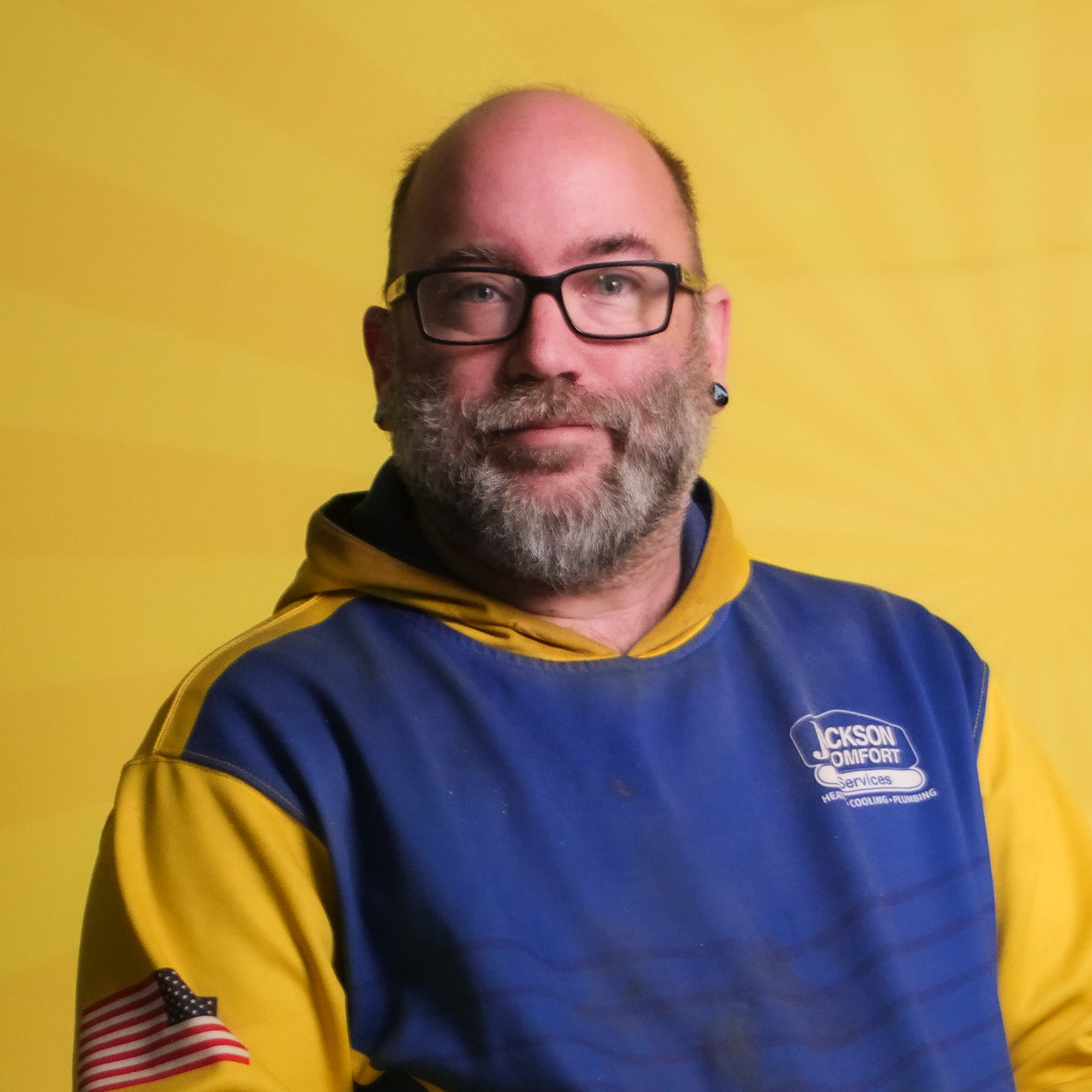 A Member Of Our Team With Glasses And A Beard Wears A Blue And Yellow Hoodie Featuring An American Flag Patch, Posing Confidently In Front Of A Yellow Background.