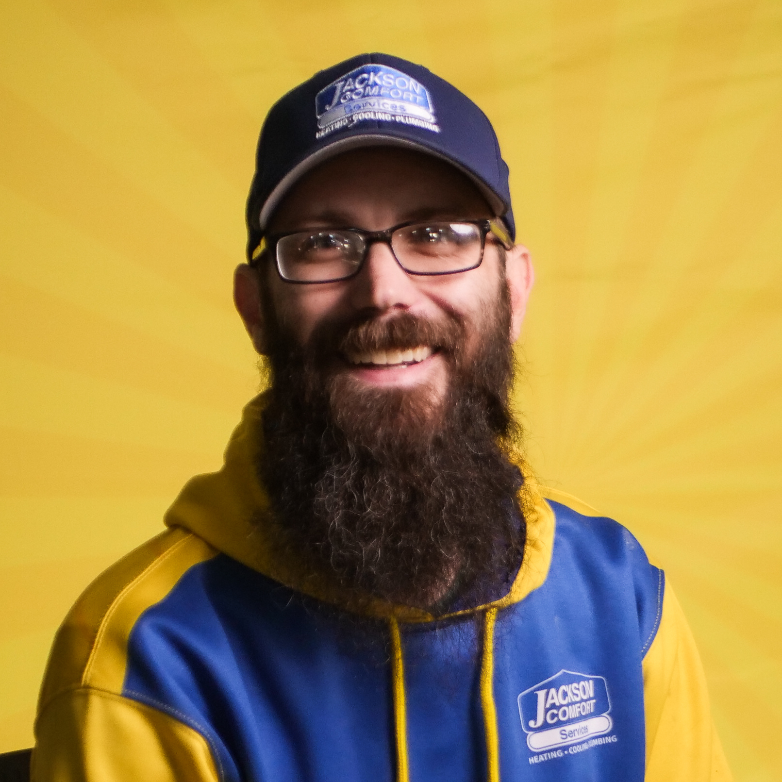 A Bearded Man Wearing Glasses, A Blue And Yellow Hoodie, And A Matching Cap Sits Smiling In Front Of A Yellow Background, Representing The Friendly Spirit Of Our Team.
