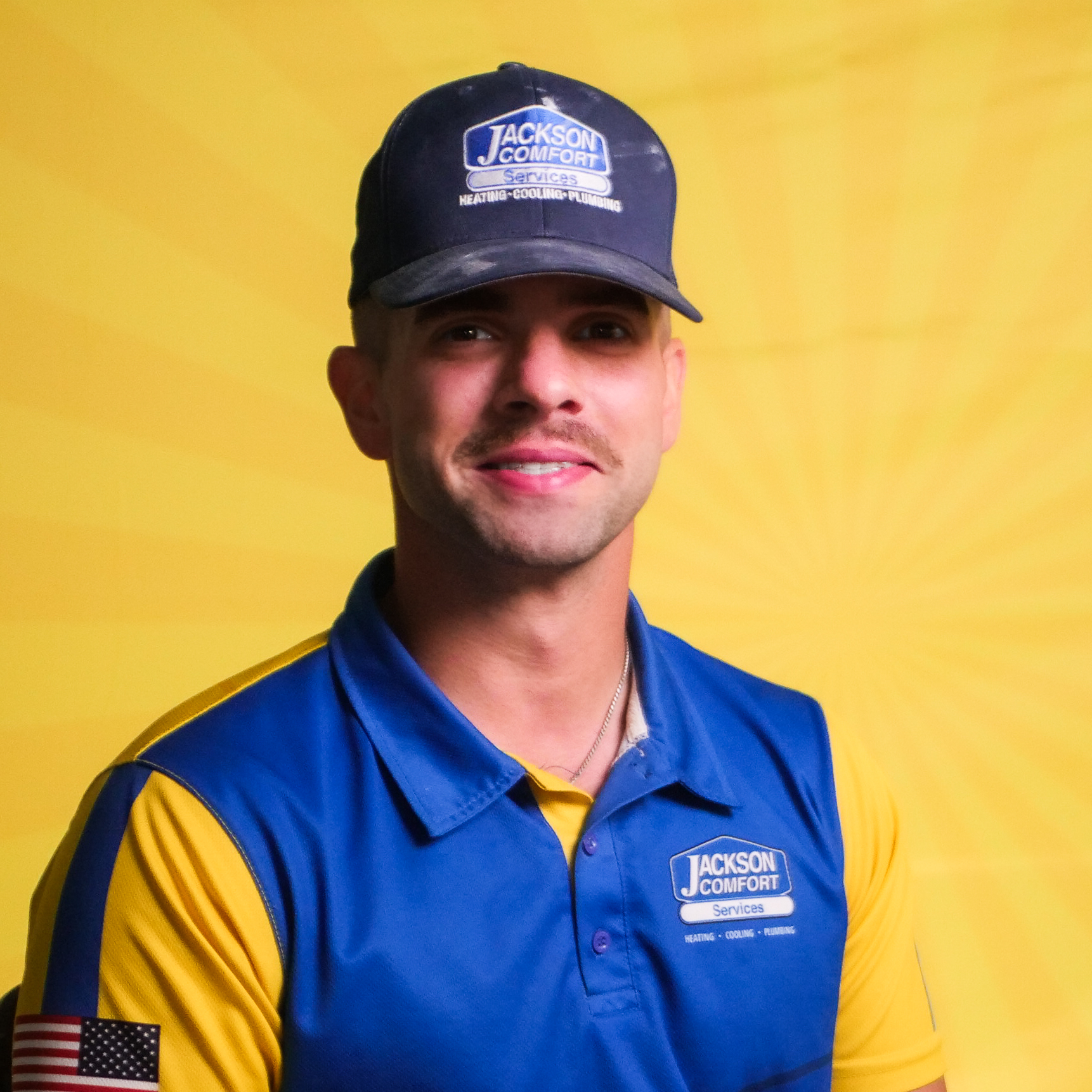 A Member Of Our Team Wears A Jackson Comfort Services Uniform And Cap As He Poses In Front Of A Yellow Background.