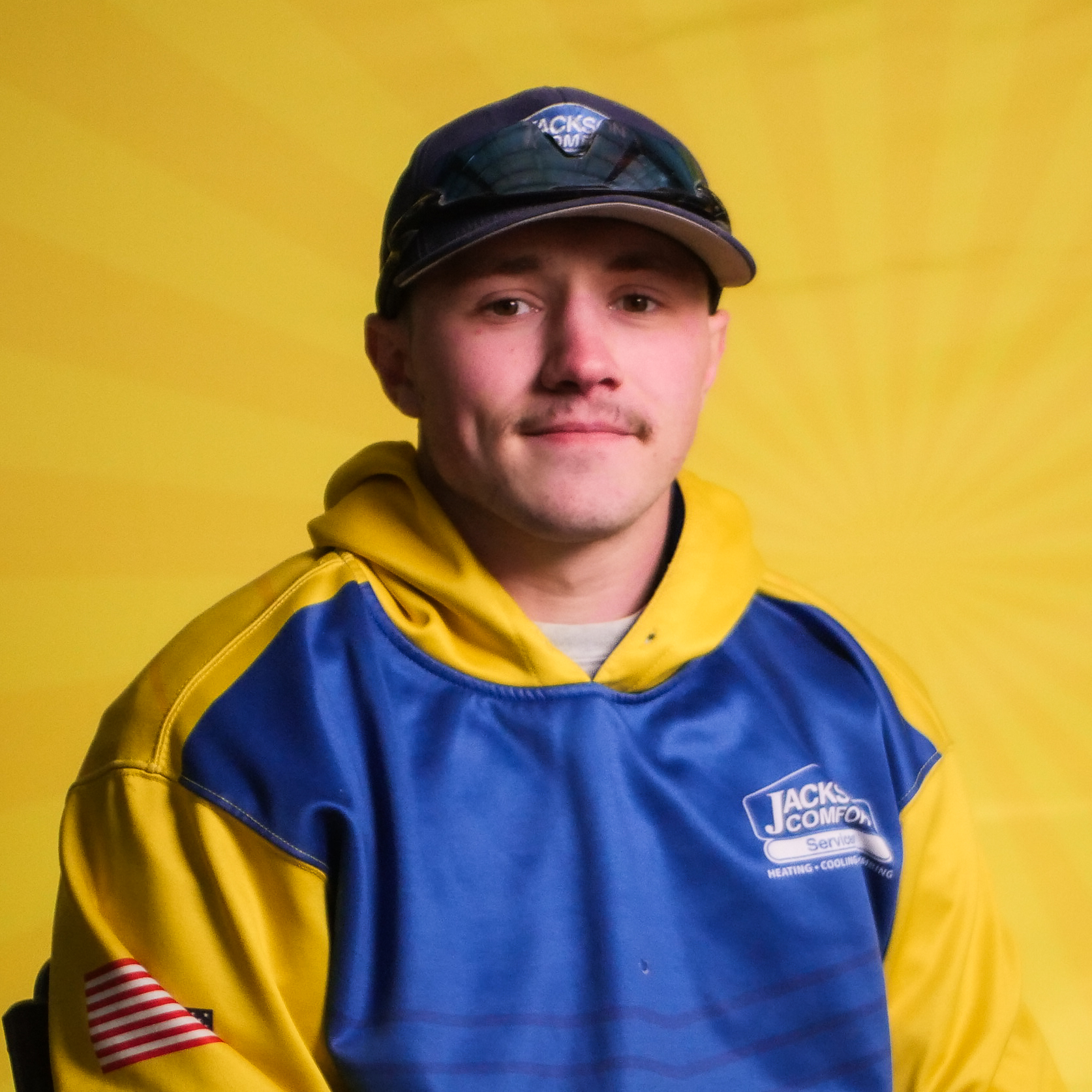 A Member Of Our Team Wearing A Blue And Yellow &Quot;Jack'S Comfort Services&Quot; Uniform And Baseball Cap Sits In Front Of A Yellow Background.