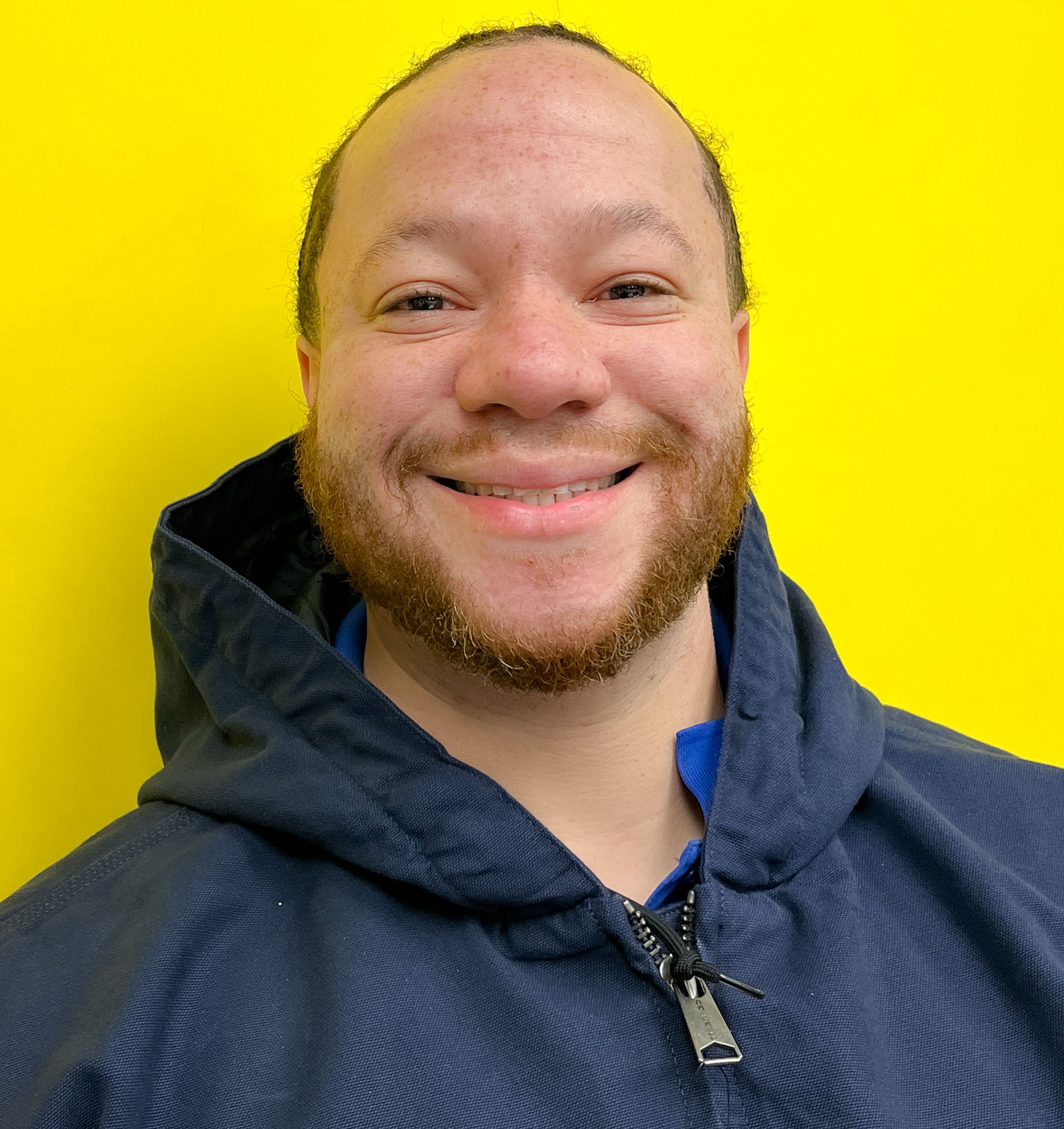 A Member Of Our Team, Wearing A Navy Blue Jacket, Smiles At The Camera In Front Of A Bright Yellow Background.