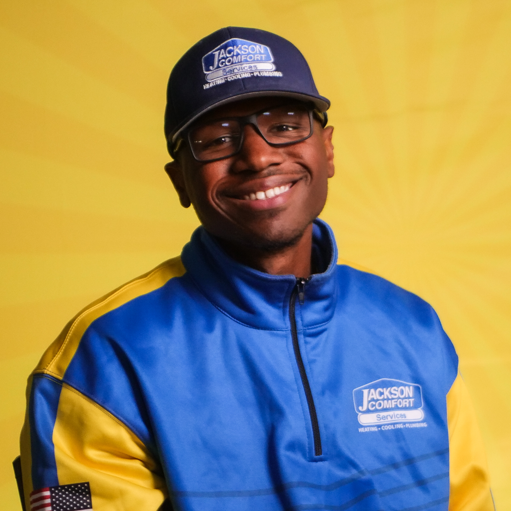 A Smiling Member Of Our Team Wears Glasses, A Blue And Yellow Jackson Comfort Services Uniform, And A Matching Cap, Posed Confidently In Front Of A Yellow Background.
