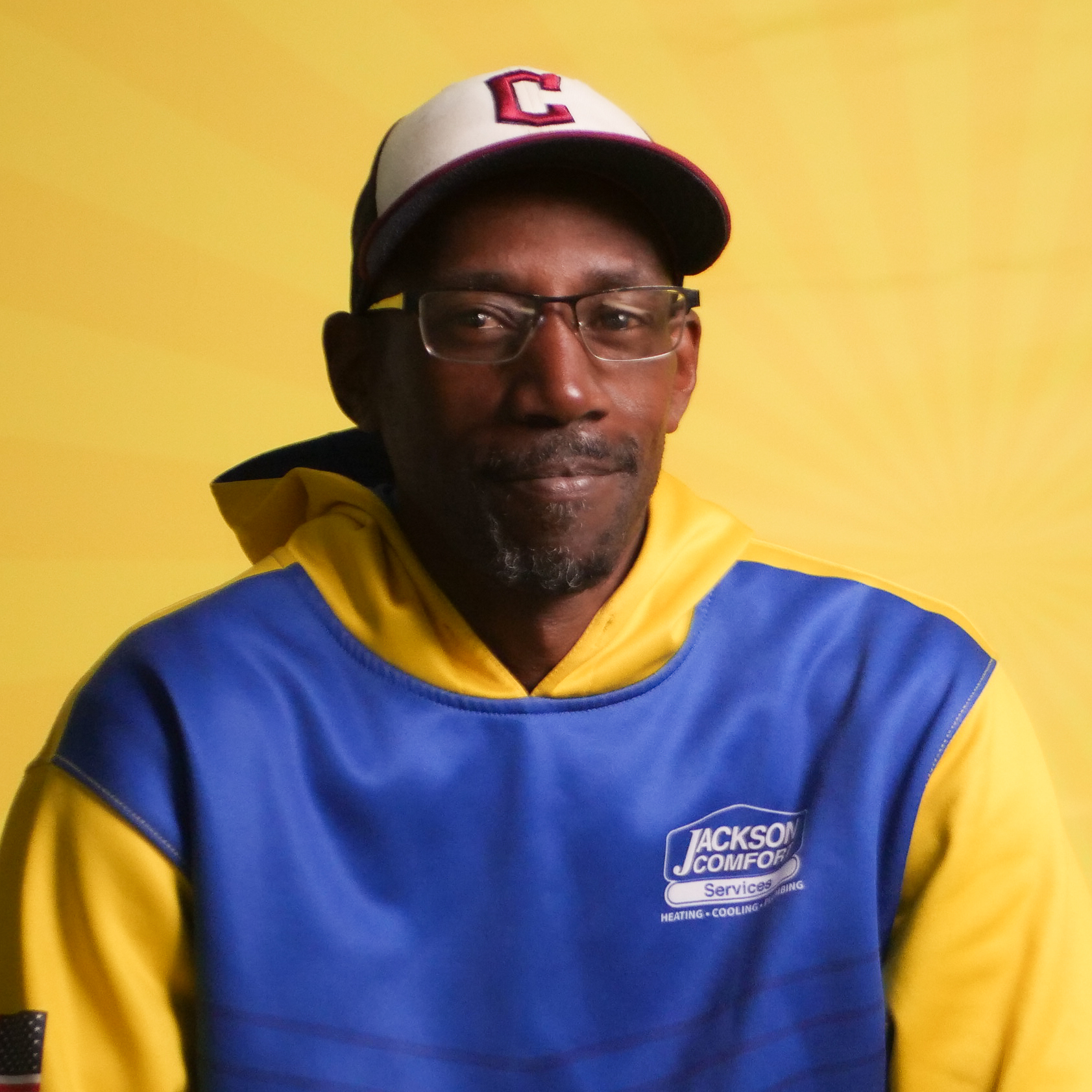A Member Of Our Team, Wearing Glasses, A Red And White Cap, And A Blue And Yellow &Quot;Jackson Comfort Services&Quot; Hoodie, Poses In Front Of A Yellow Background.