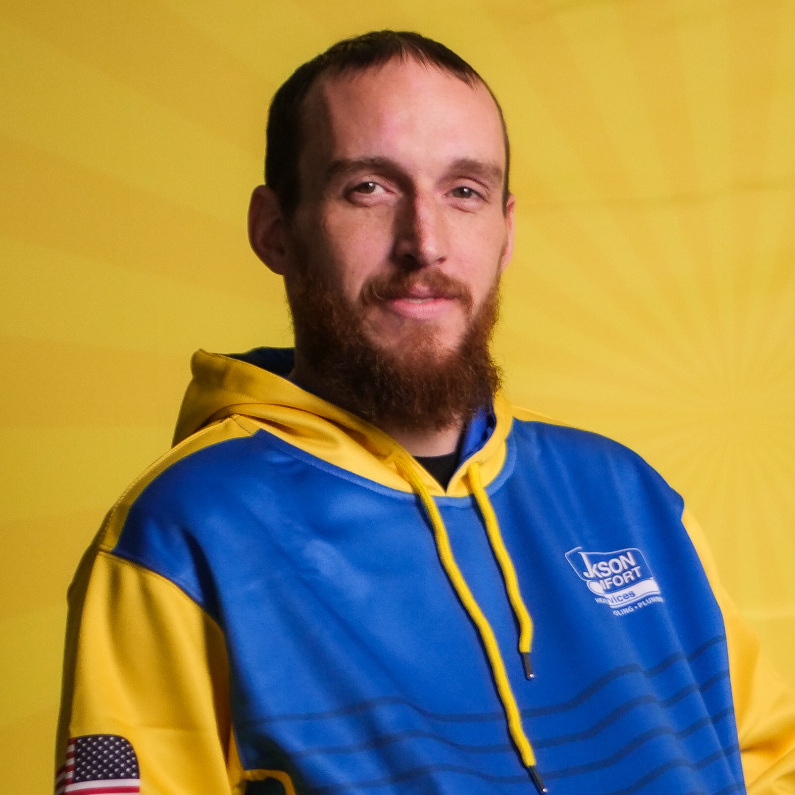 A Member Of Our Team With A Beard, Wearing A Blue And Yellow Hoodie, Stands In Front Of A Yellow Background With A Sunburst Pattern.