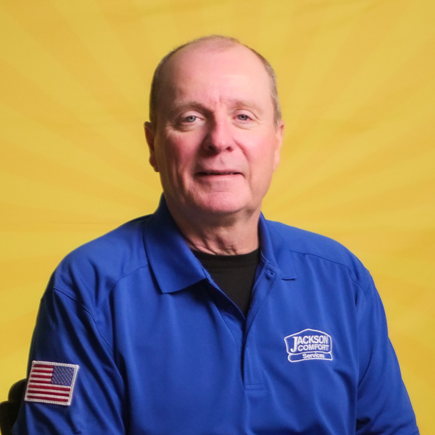 A Member Of Our Team In A Blue Jackson Comfort Services Shirt With An American Flag Patch On The Sleeve Poses In Front Of A Bright Yellow Background.