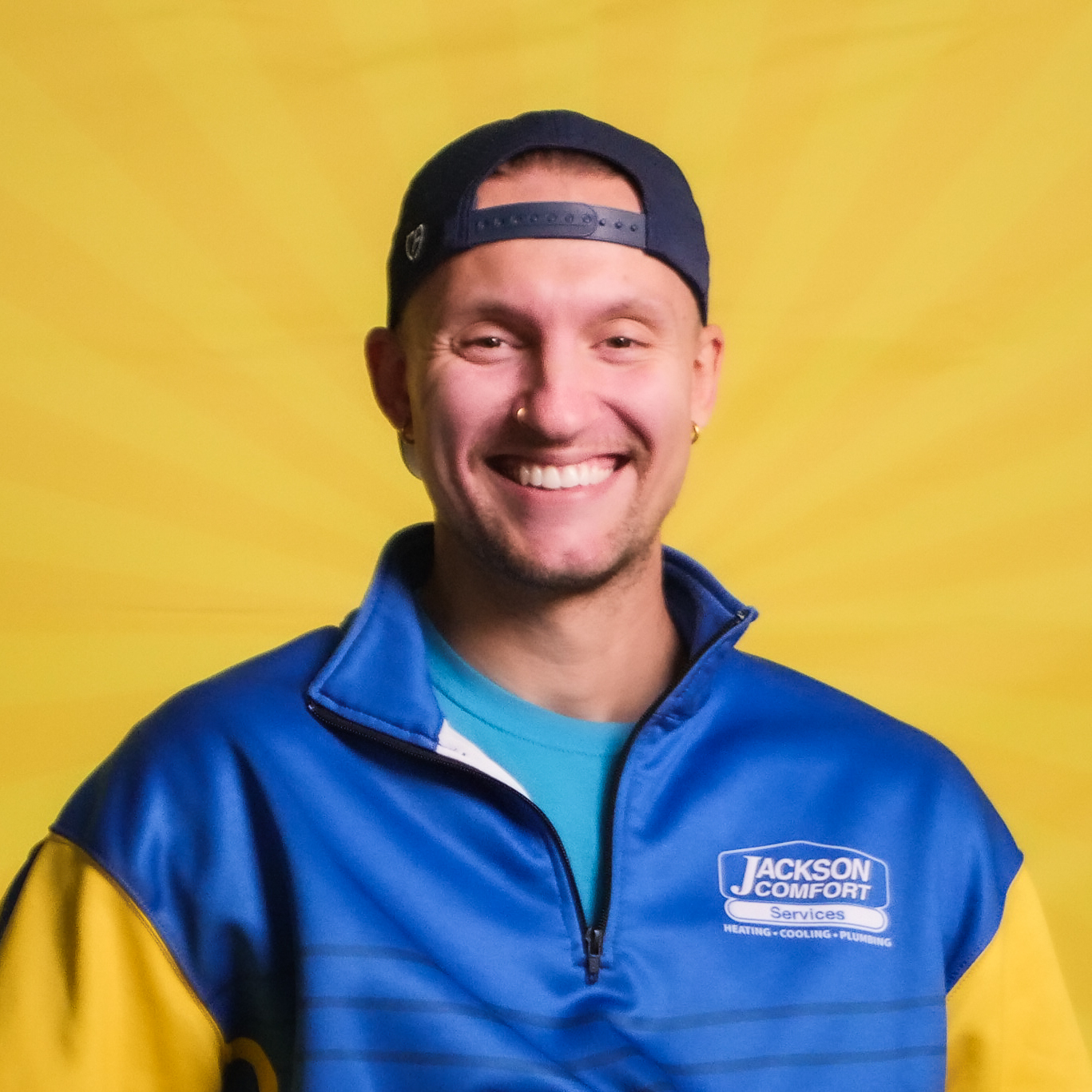 A Smiling Member Of Our Team In A Blue &Quot;Jackson Comfort Services&Quot; Jacket And Backwards Cap Stands In Front Of A Yellow Background.