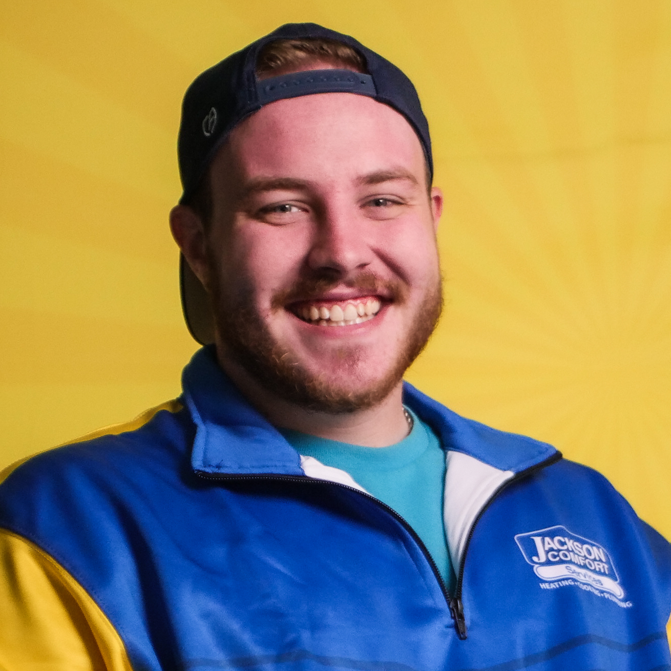 A Man Wearing A Blue And Yellow Jacket And A Backwards Cap Is Smiling In Front Of A Yellow Background, Representing The Positive Spirit Of Our Team.
