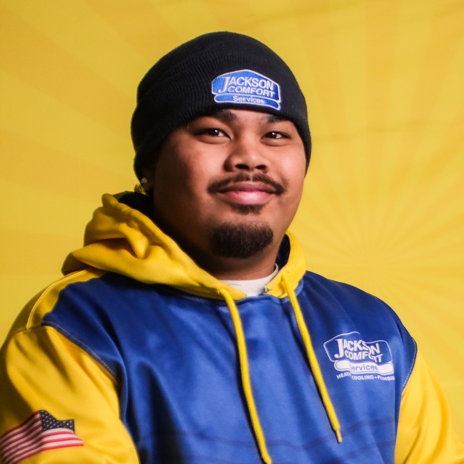 A Member Of Our Team, Wearing A Blue And Yellow Jackson Comfort Service Uniform And Beanie, Smiles In Front Of A Yellow Background.