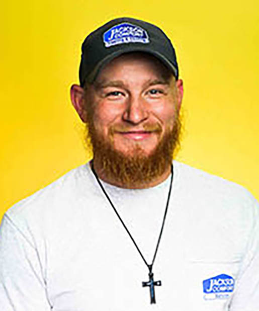 A Man With A Red Beard, Part Of Our Team, Wears A Black Cap, A White Shirt With A Blue Logo, And A Black Cross Necklace As He Stands In Front Of A Yellow Background.