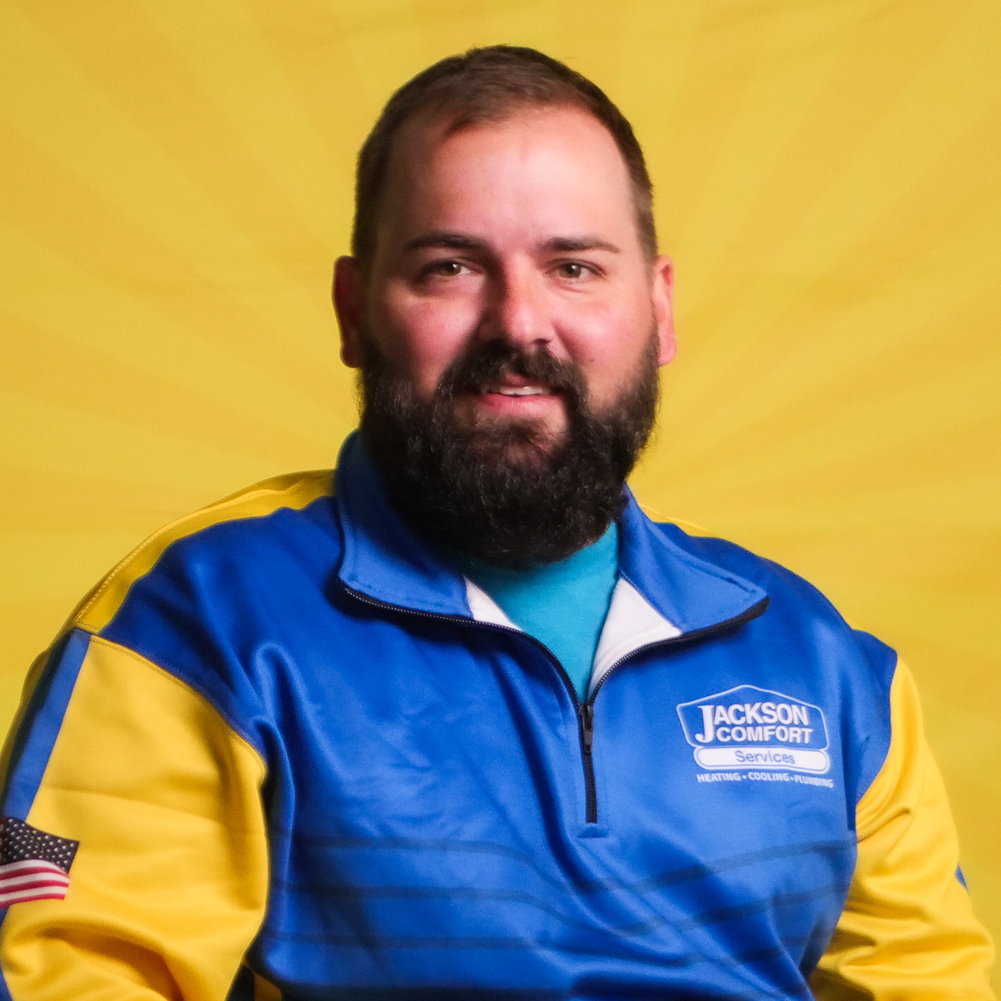 A Bearded Man From Our Team Wears A Blue And Yellow &Quot;Jackson Comfort Services&Quot; Jacket As He Poses Confidently In Front Of A Vibrant Yellow Background.