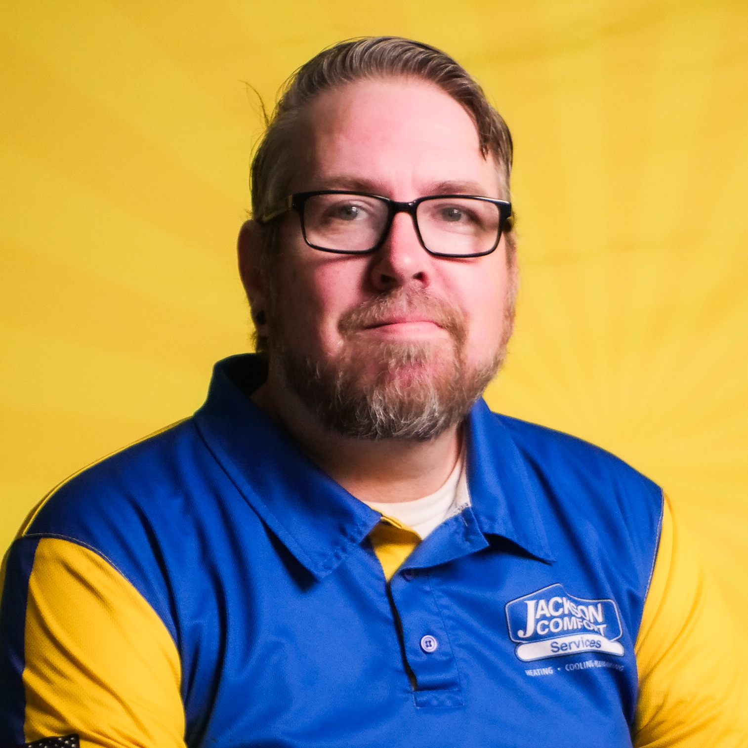 A Man With Glasses And A Beard, Representing Our Team, Wears A Blue And Yellow Jackson Comfort Services Shirt As He Sits In Front Of A Yellow Background.