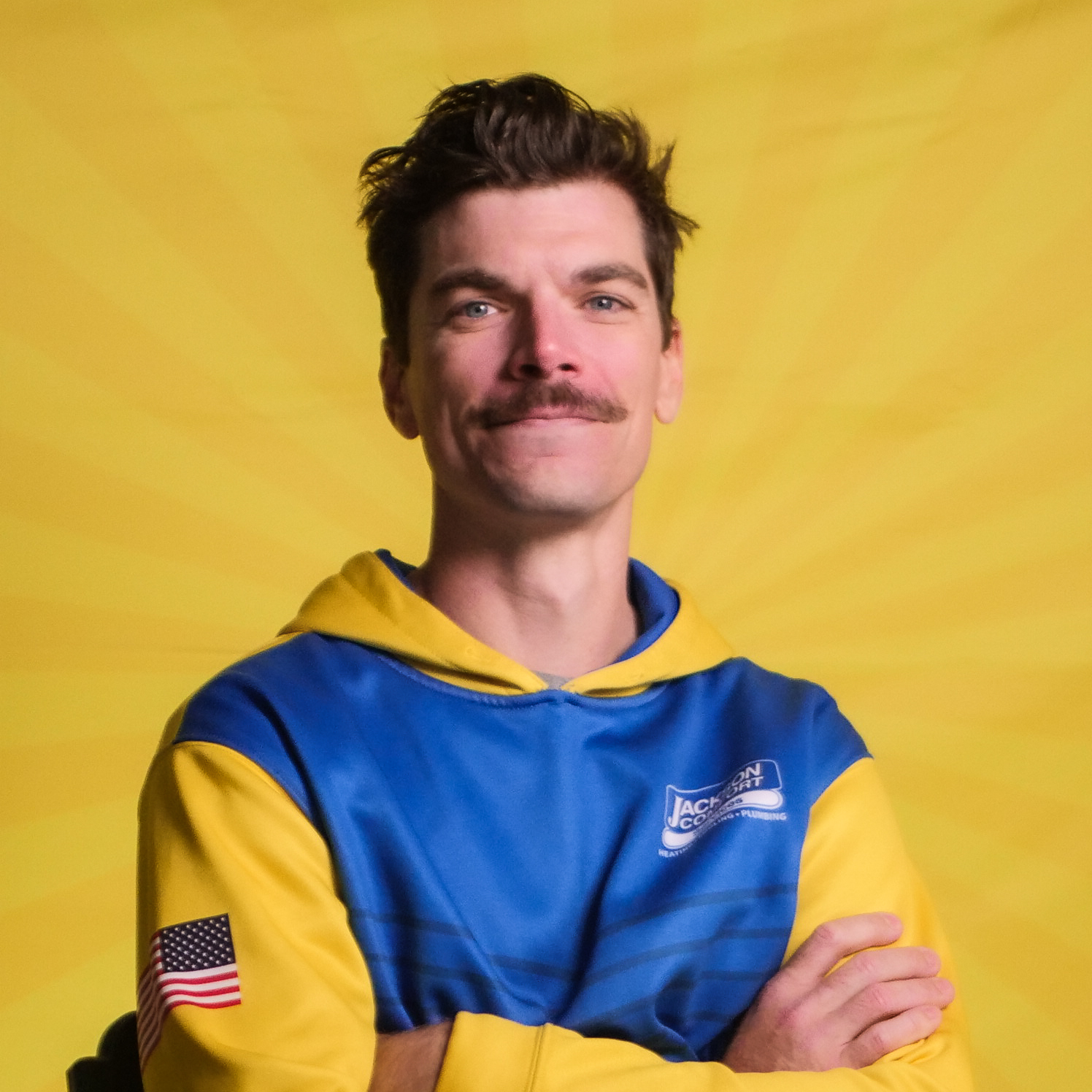 A Man With A Mustache Wearing A Blue And Yellow Hoodie Stands With Arms Crossed In Front Of A Yellow Background With Radiating Lines, Representing The Confident Spirit Of Our Team.