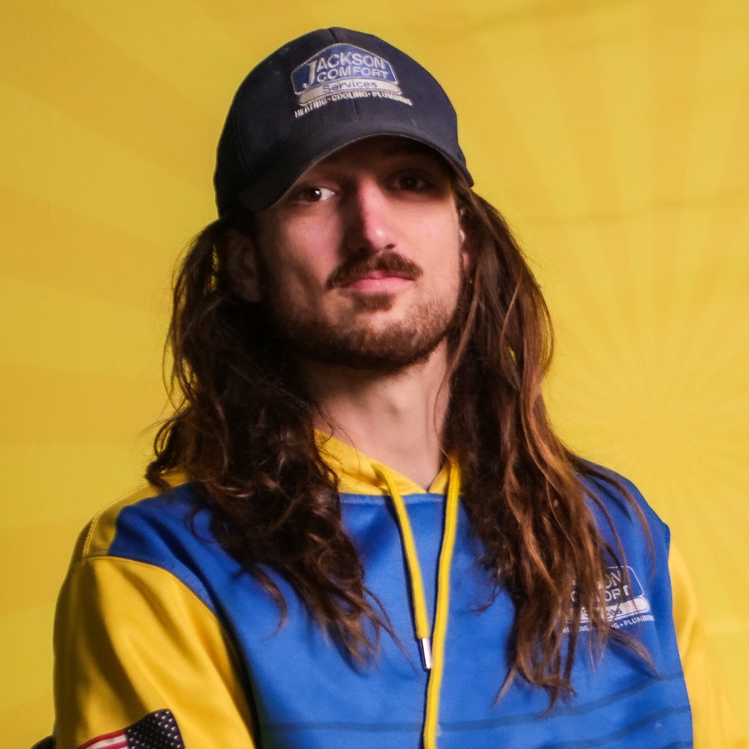 A Member Of Our Team With Long Hair And A Beard Wears A Blue And Yellow Hoodie And A Jackson Comfort Cap, Standing In Front Of A Yellow Background With A Subtle Radiating Pattern.