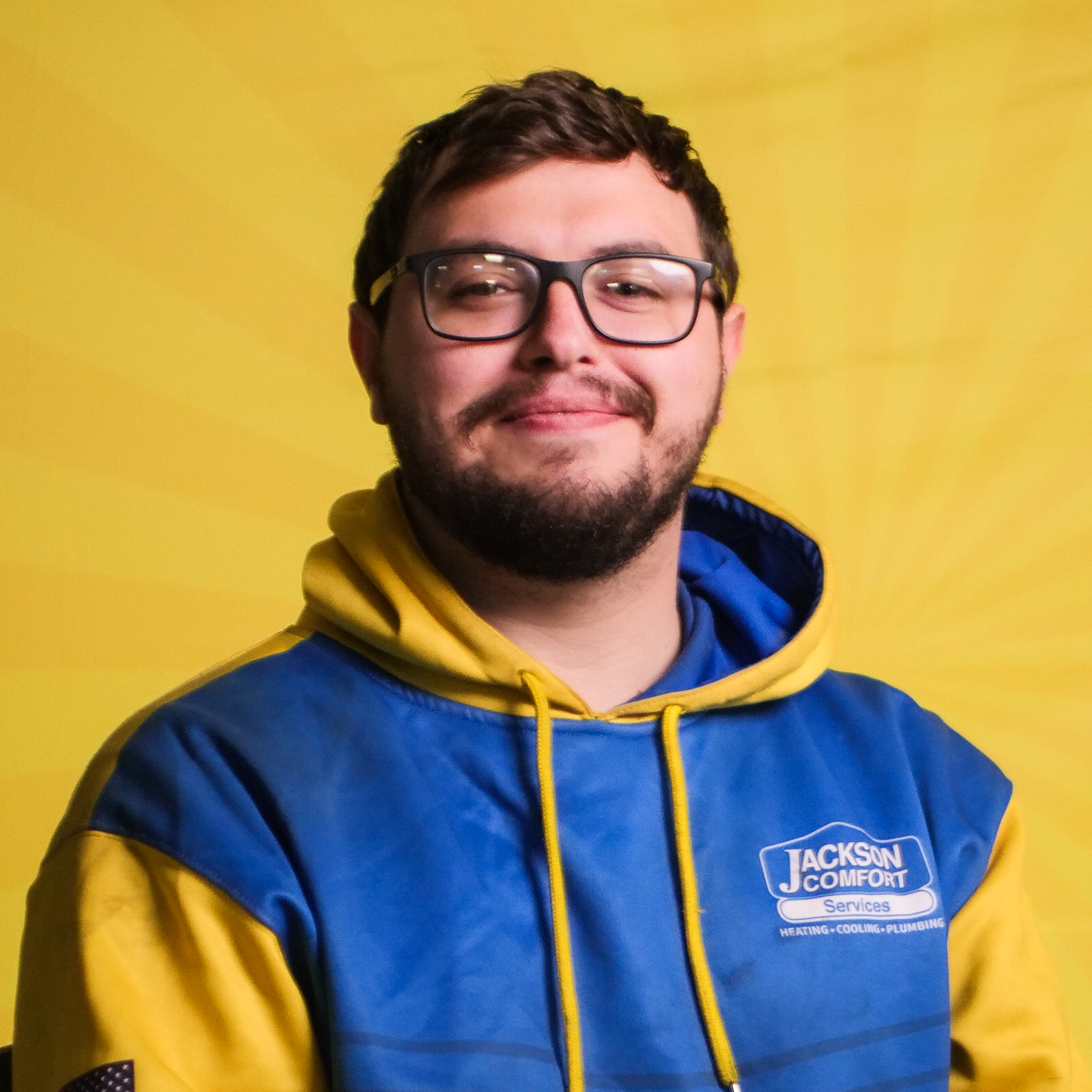 A Member Of Our Team, Wearing Glasses And A Blue And Yellow Jackson Comfort Services Hoodie, Poses Confidently In Front Of A Yellow Background.