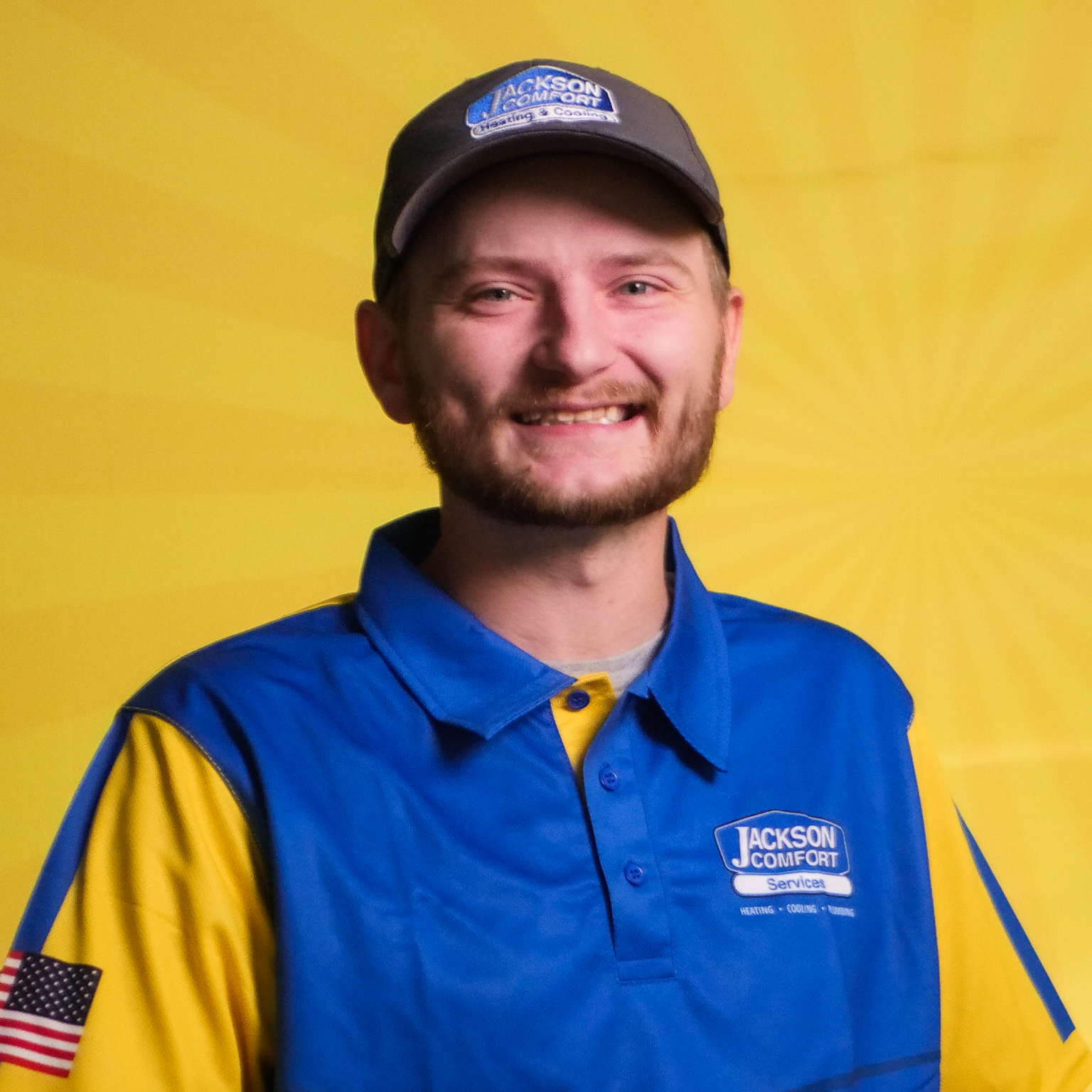 A Smiling Member Of Our Team, Wearing A Blue And Yellow Jackson Comfort Services Uniform And Cap, Stands In Front Of A Yellow Background.