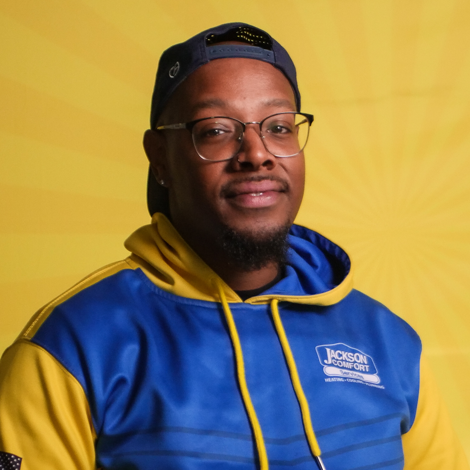 A Member Of Our Team Wears Glasses, A Blue And Yellow &Quot;Jackson Comfort&Quot; Hoodie, And A Backwards Cap While Standing In Front Of A Vibrant Yellow Background.