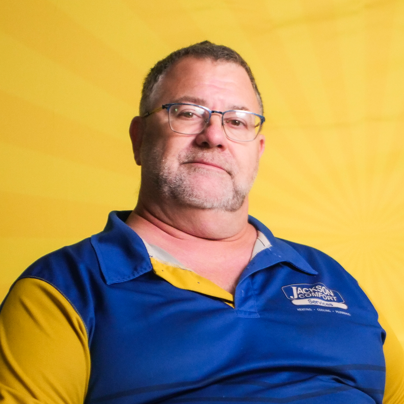 Middle-Aged Man With Glasses And A Trimmed Beard, Wearing A Blue And Yellow Collared Shirt, Sits In Front Of A Yellow Background With Radiating Lines, Representing A Valued Member Of Our Team.