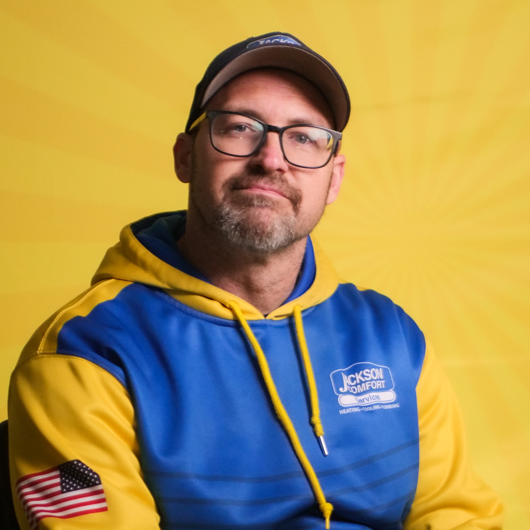 A Man Wearing Glasses, A Cap, And A Blue And Yellow Jackson Comfort Hoodie With An American Flag Patch On The Sleeve Sits Against A Yellow Background, Representing Our Team.