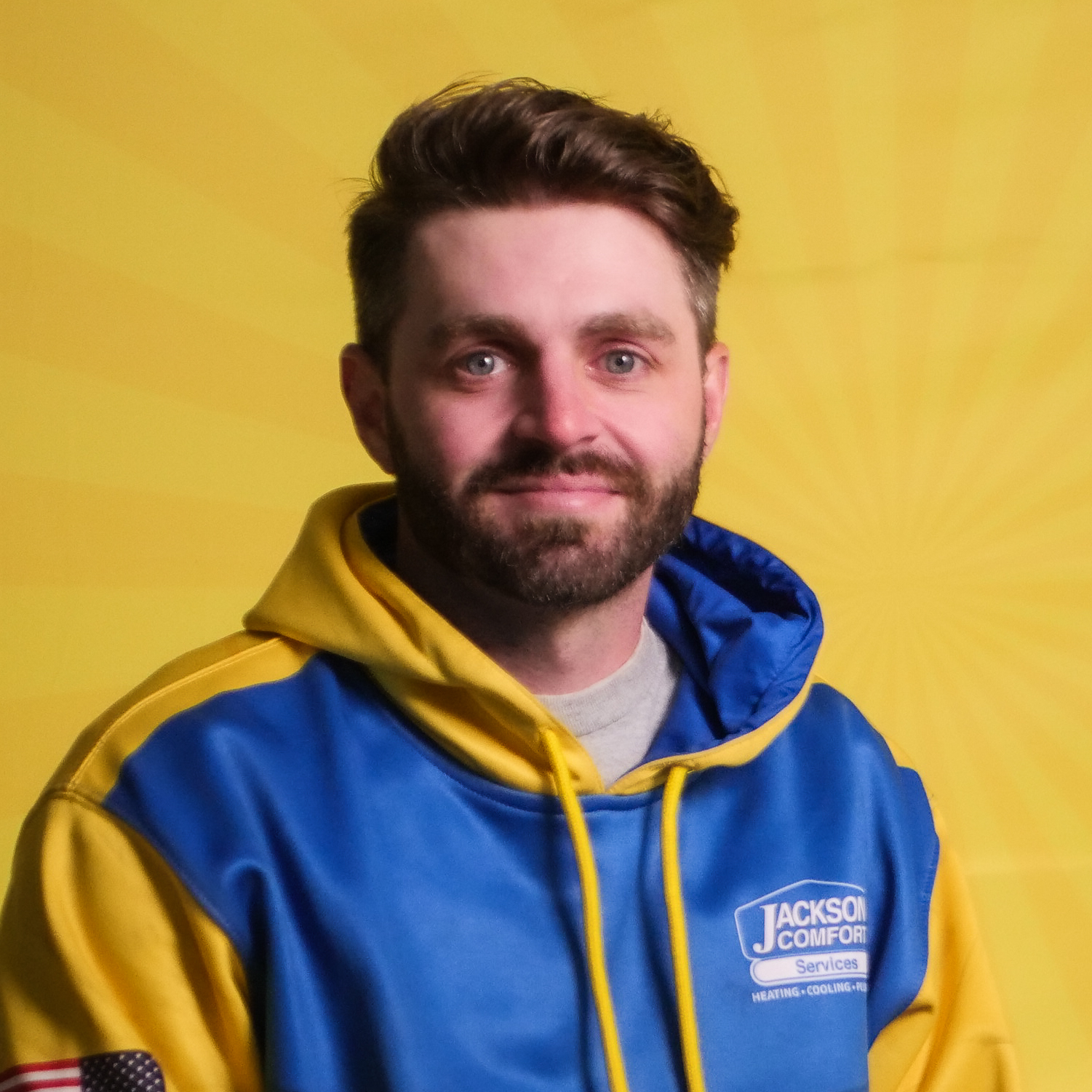 A Man With Brown Hair And A Beard, Representing Our Team, Wears A Blue And Yellow Jackson Comfort Services Hoodie As He Poses In Front Of A Yellow Background With A Sunburst Pattern.