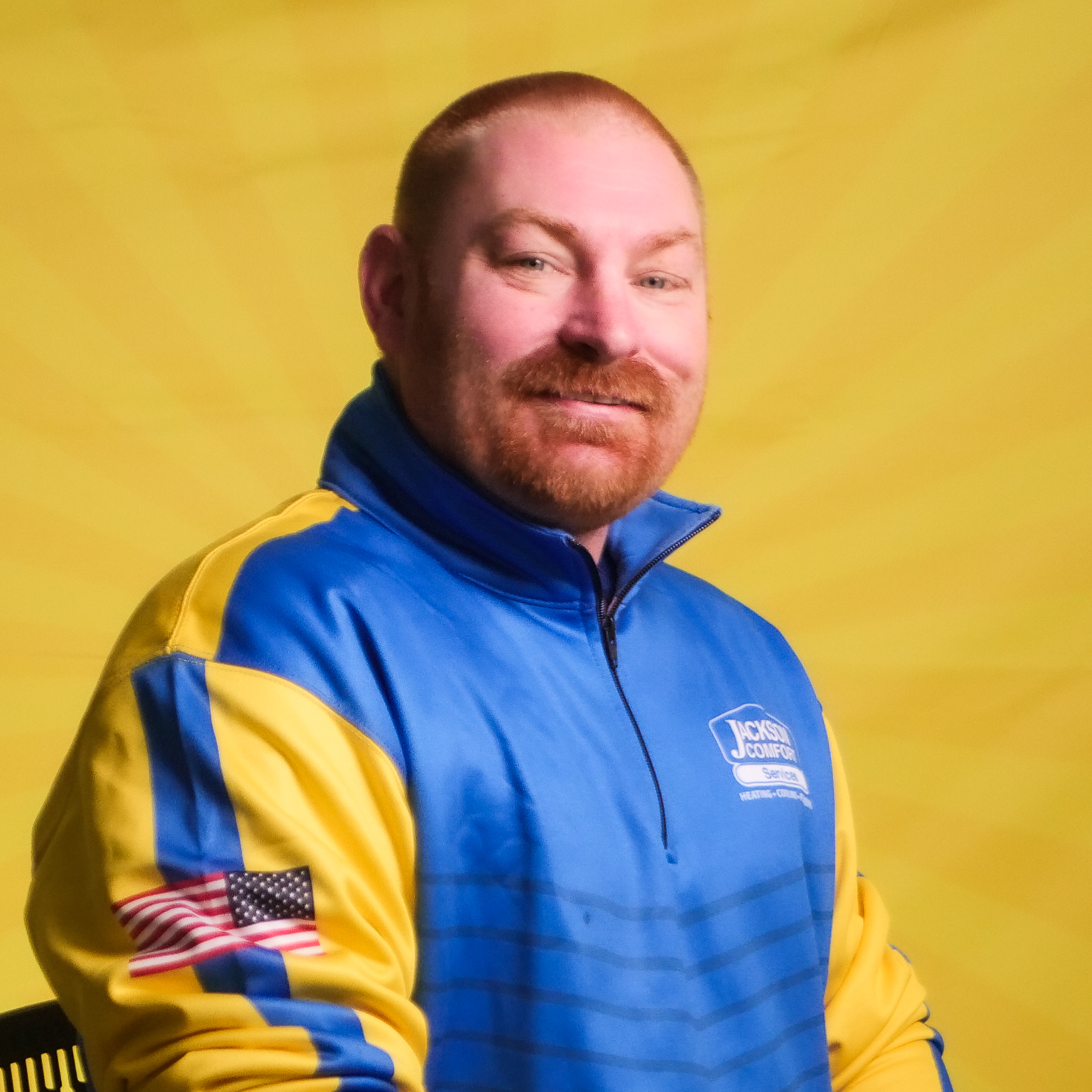 A Member Of Our Team, A Man With A Red Beard In A Blue And Yellow Jacket Featuring An American Flag Patch, Sits In Front Of A Yellow Background.