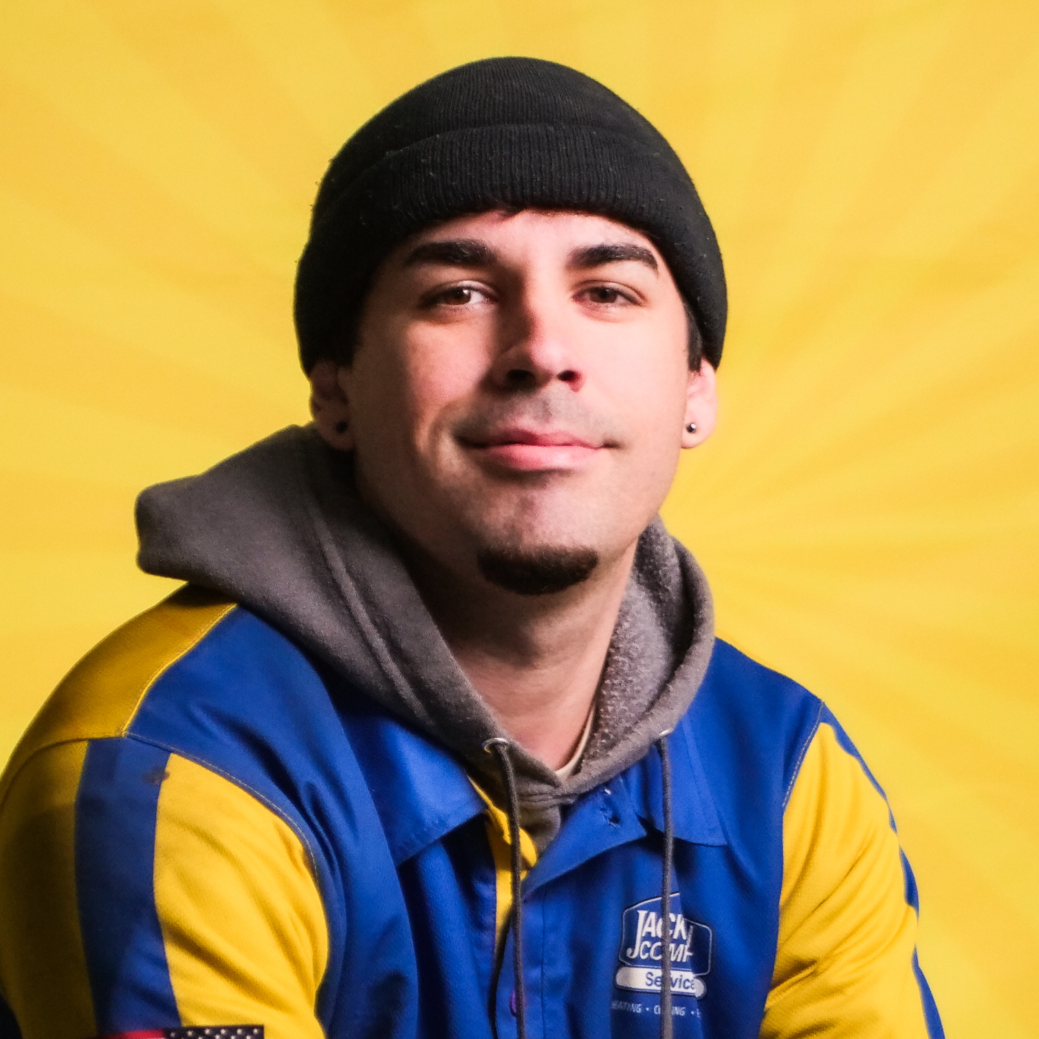 A Member Of Our Team, Wearing A Black Beanie And A Blue-And-Yellow Jacket, Sits In Front Of A Bright Yellow Background, Looking At The Camera.