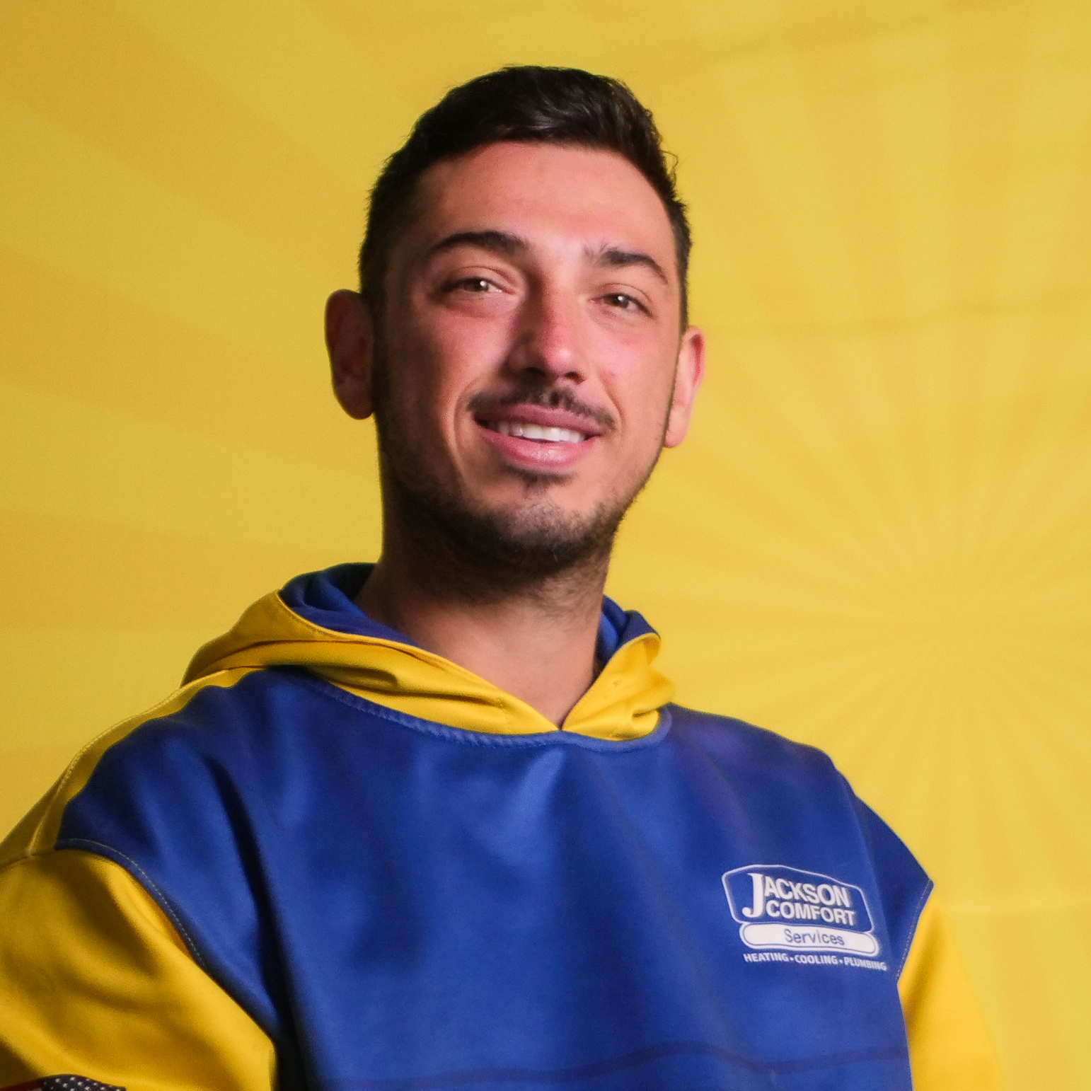 A Man Wearing A Blue And Yellow Jackson Comfort Services Hoodie, Representing Our Team, Poses In Front Of A Yellow Background With A Subtle Radiating Pattern.
