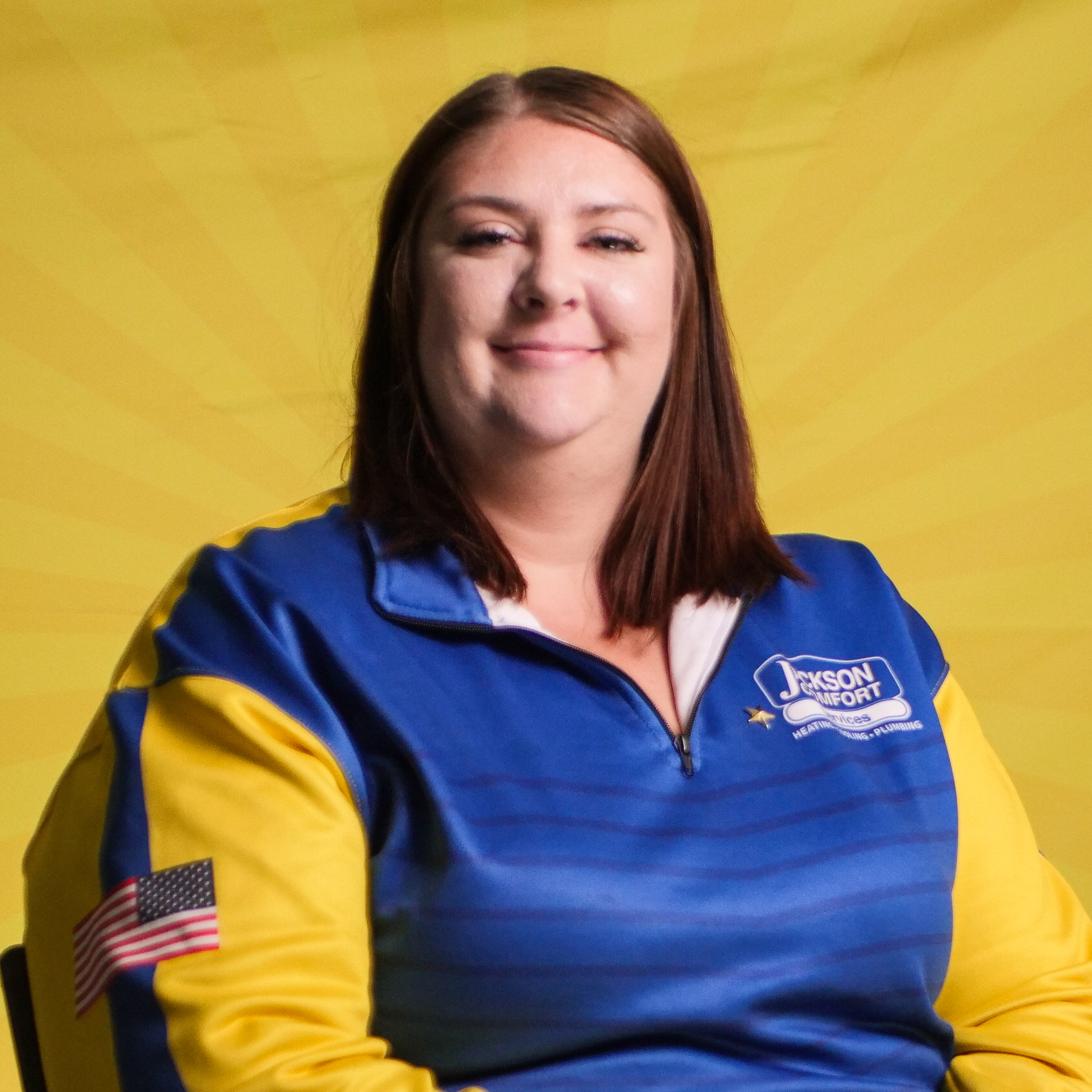 A Member Of Our Team With Straight Brown Hair, Wearing A Blue And Yellow Jacket With An American Flag Patch, Sits In Front Of A Yellow Background Featuring A Subtle Sunburst Pattern.