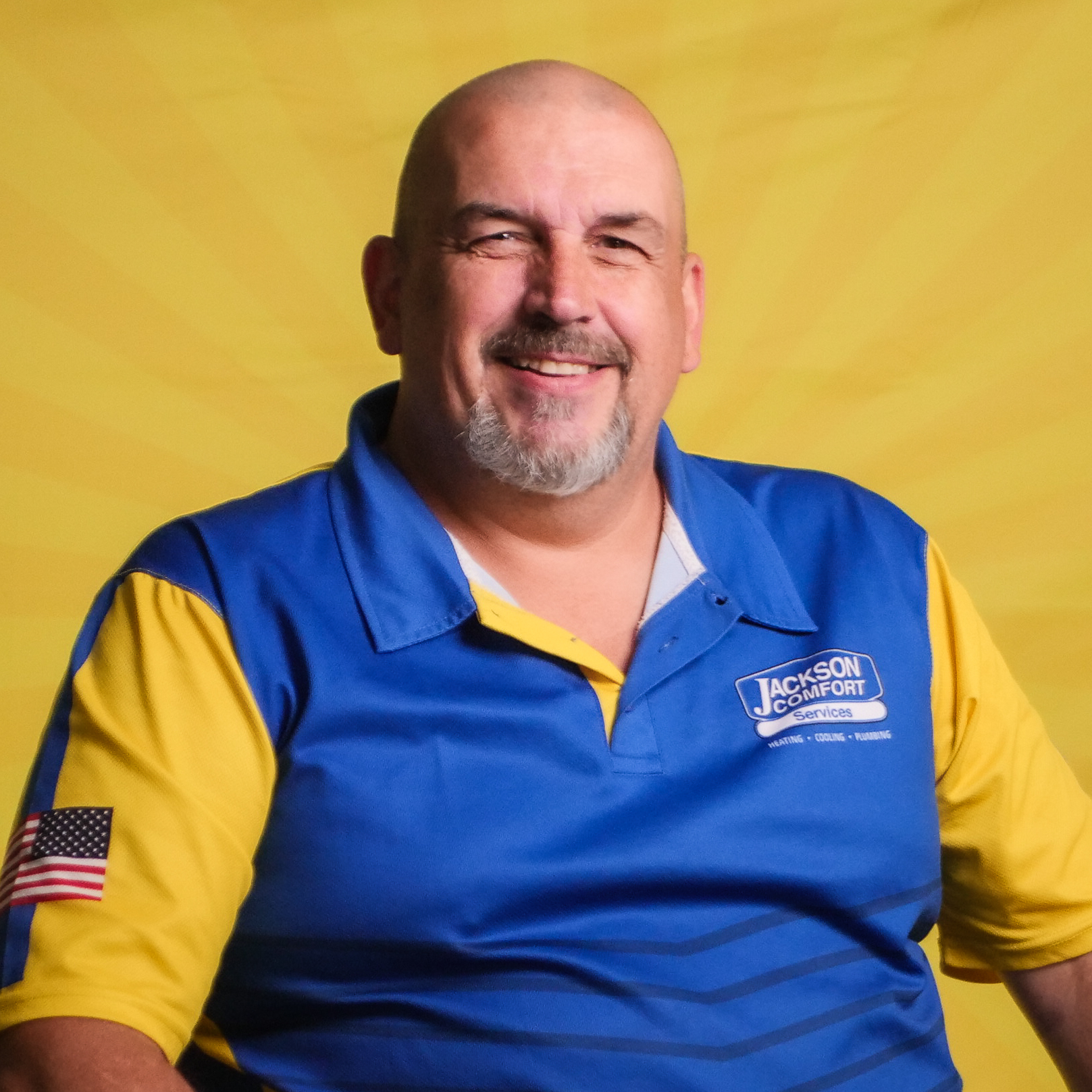 Bald Man With A Goatee Smiling, Wearing A Blue And Yellow Jackson Comfort Services Polo Shirt With A Us Flag Patch On The Sleeve, Seated In Front Of A Yellow Background—Proudly Representing Our Team.