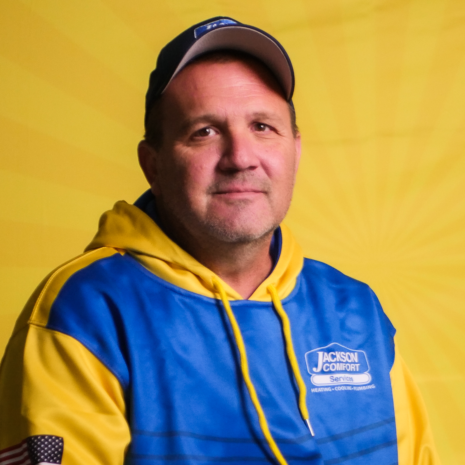 A Member Of Our Team In A Blue And Yellow &Quot;Jackson Comfort&Quot; Hoodie And Cap Sits In Front Of A Yellow Background With A Subtle Radiating Pattern.