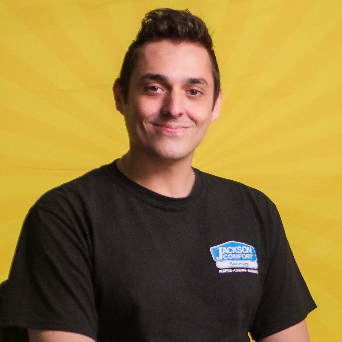 A Member Of Our Team Wears A Black Jackson Comfort T-Shirt While Sitting In Front Of A Yellow Background With A Subtle Sunburst Pattern.