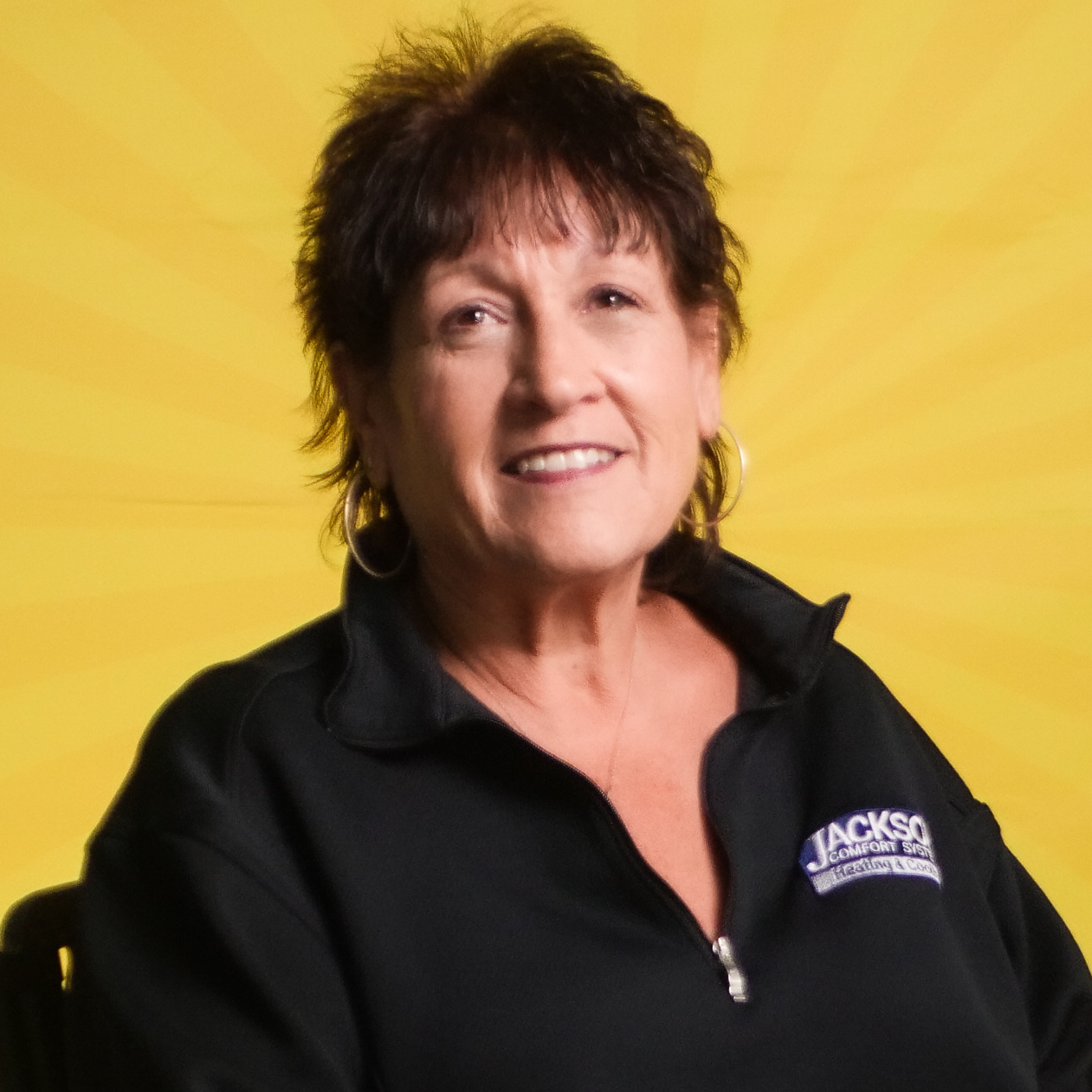 A Woman With Short Brown Hair, Wearing A Black Zip-Up Jacket With A &Quot;Jackson&Quot; Logo, Sits Smiling In Front Of A Yellow Sunburst Background—Proudly Representing Our Team.