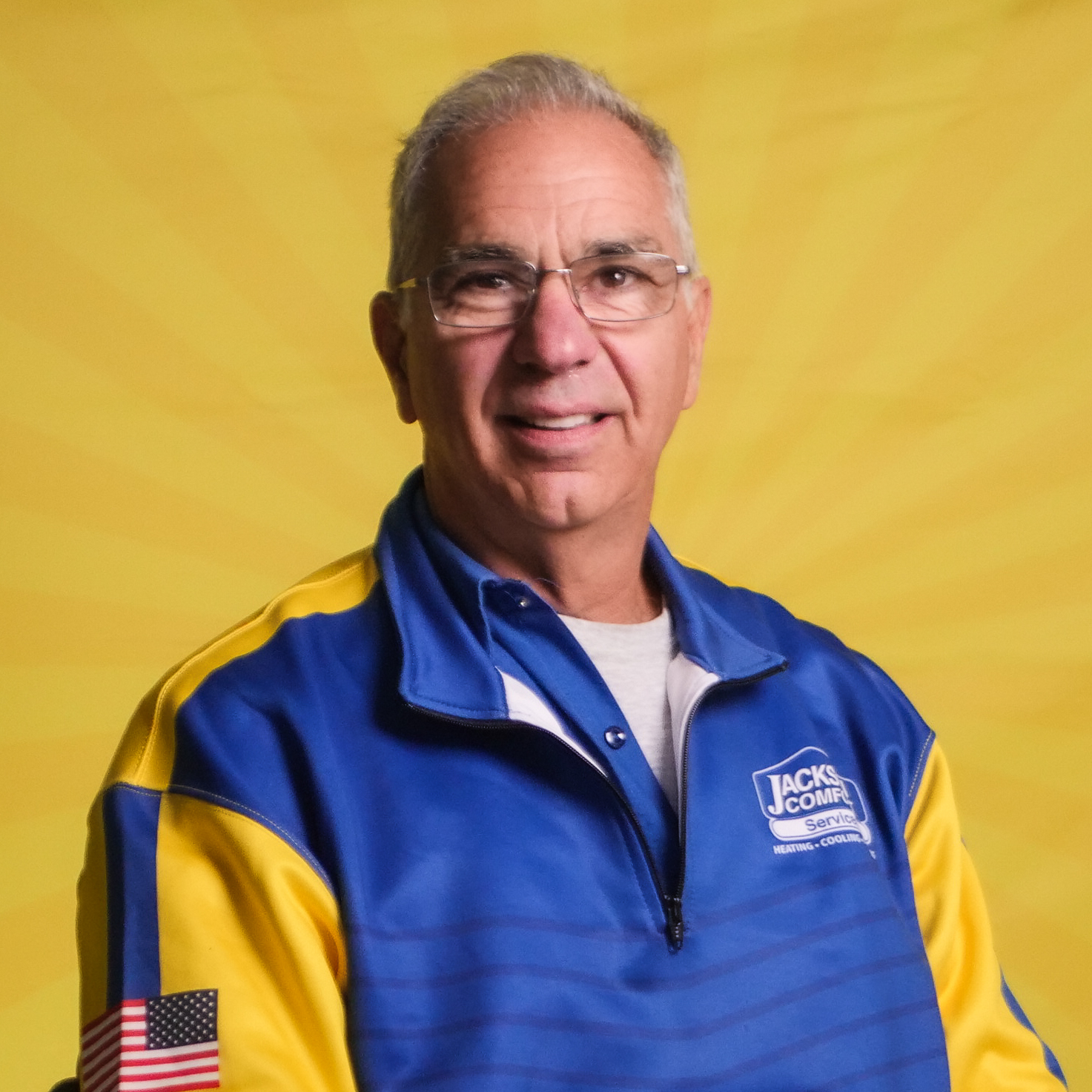 An Older Man Wearing Glasses And A Blue And Yellow &Quot;Jack'S Complete&Quot; Jacket Stands In Front Of A Yellow Background, Representing Our Team With Pride.