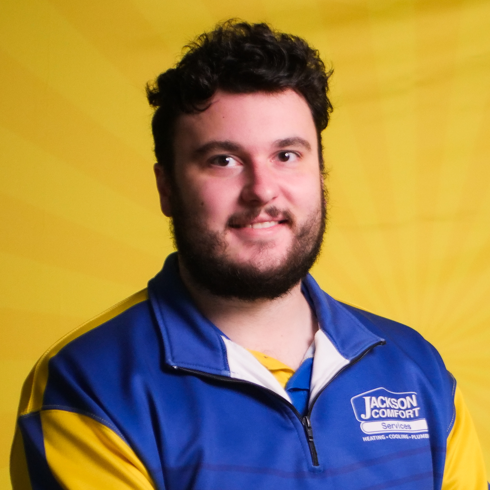 A Man With Dark Hair And A Beard, Part Of Our Team, Wears A Blue And Yellow Jackson Comfort Services Jacket, Standing In Front Of A Yellow Background.