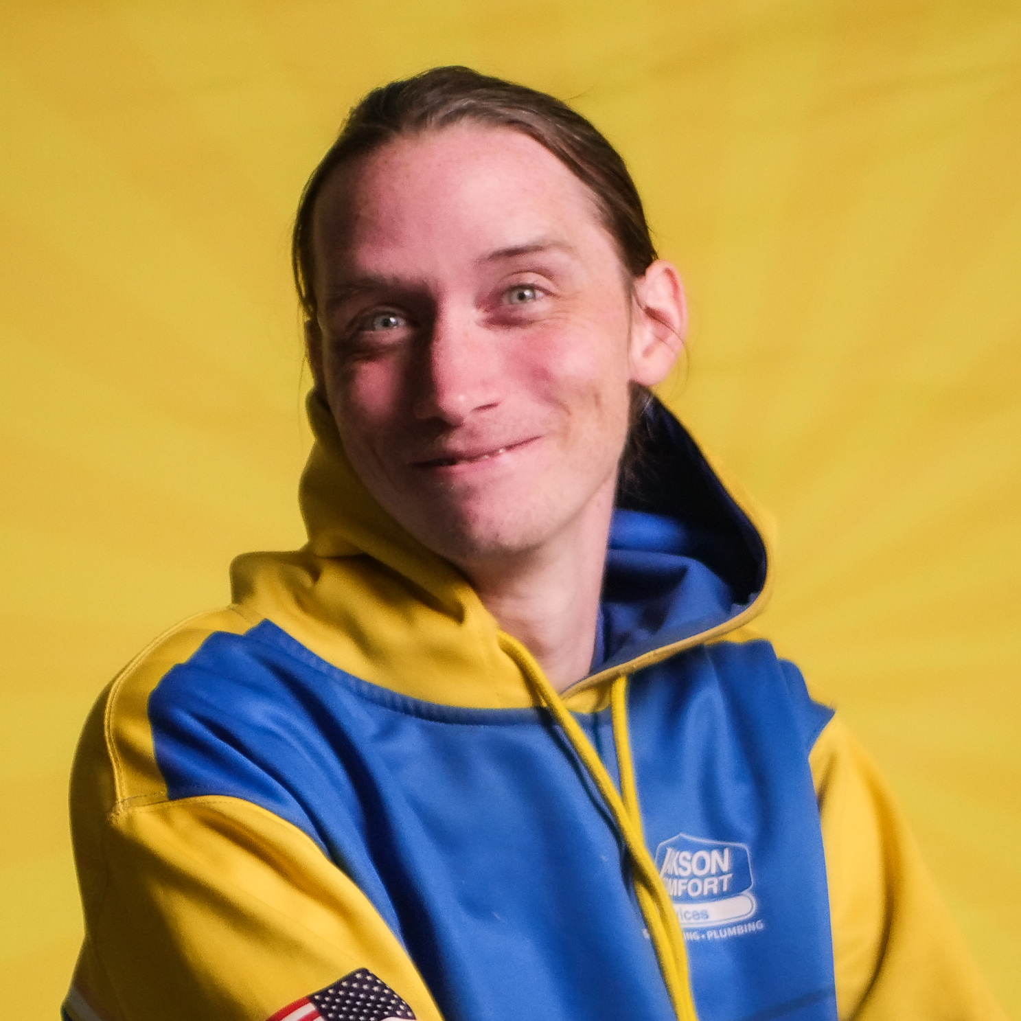 A Member Of Our Team With Long Hair In A Yellow And Blue Hooded Jacket, Featuring An American Flag Patch, Smiles In Front Of A Bright Yellow Background.