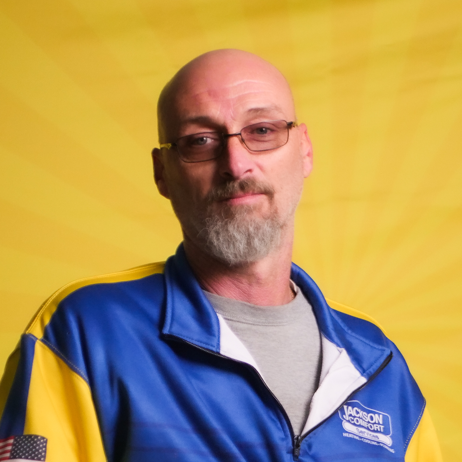 A Man With Glasses, A Bald Head, And A Gray Goatee Wears A Blue And Yellow Jackson Comfort Jacket, Representing Our Team As He Stands In Front Of A Yellow Background.