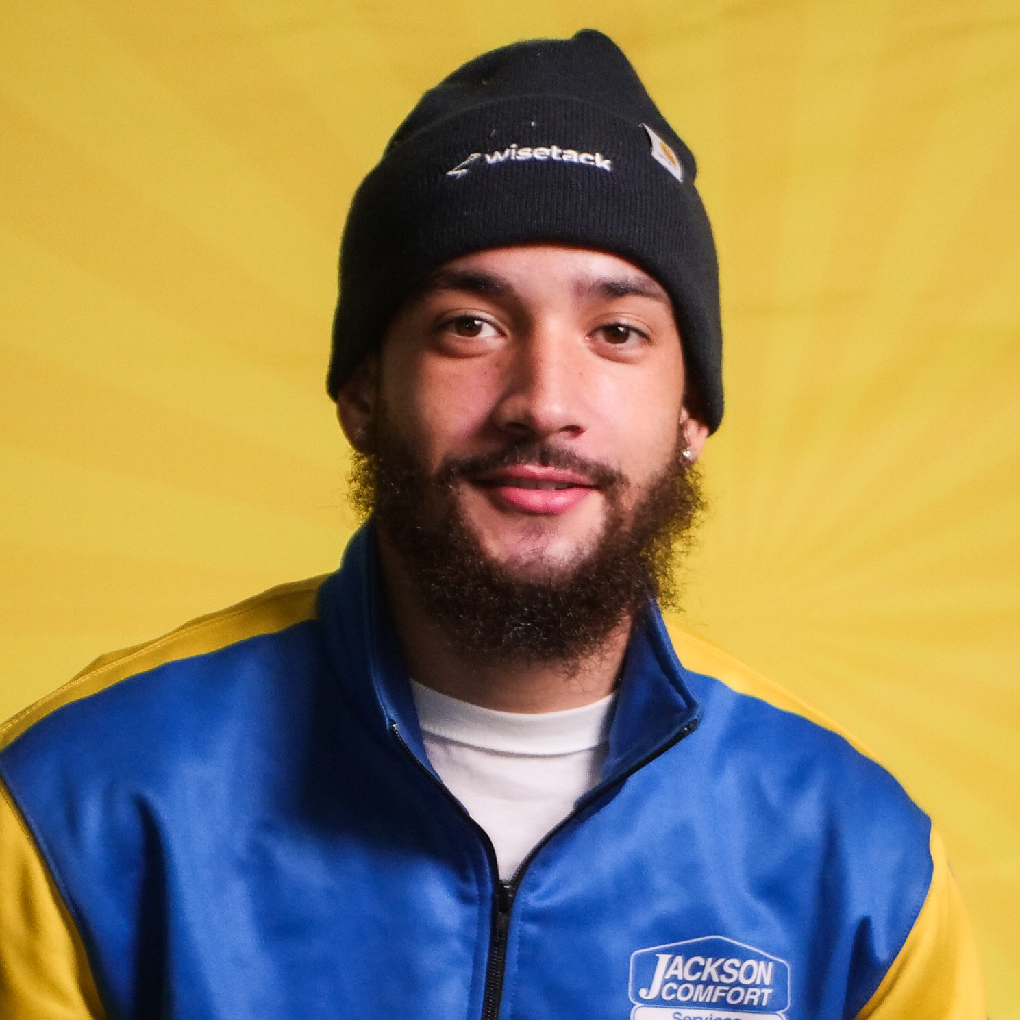 A Member Of Our Team With A Beard Wears A Black Beanie, Blue And Yellow &Quot;Jackson Comfort&Quot; Jacket, And White Shirt While Sitting In Front Of A Bright Yellow Background.