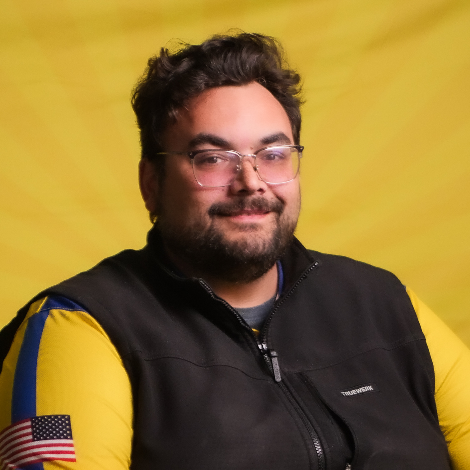 A Member Of Our Team, A Man With Glasses And A Beard, Wears A Black Vest And A Yellow Shirt With An American Flag Patch On The Sleeve, Sitting In Front Of A Bright Yellow Background.