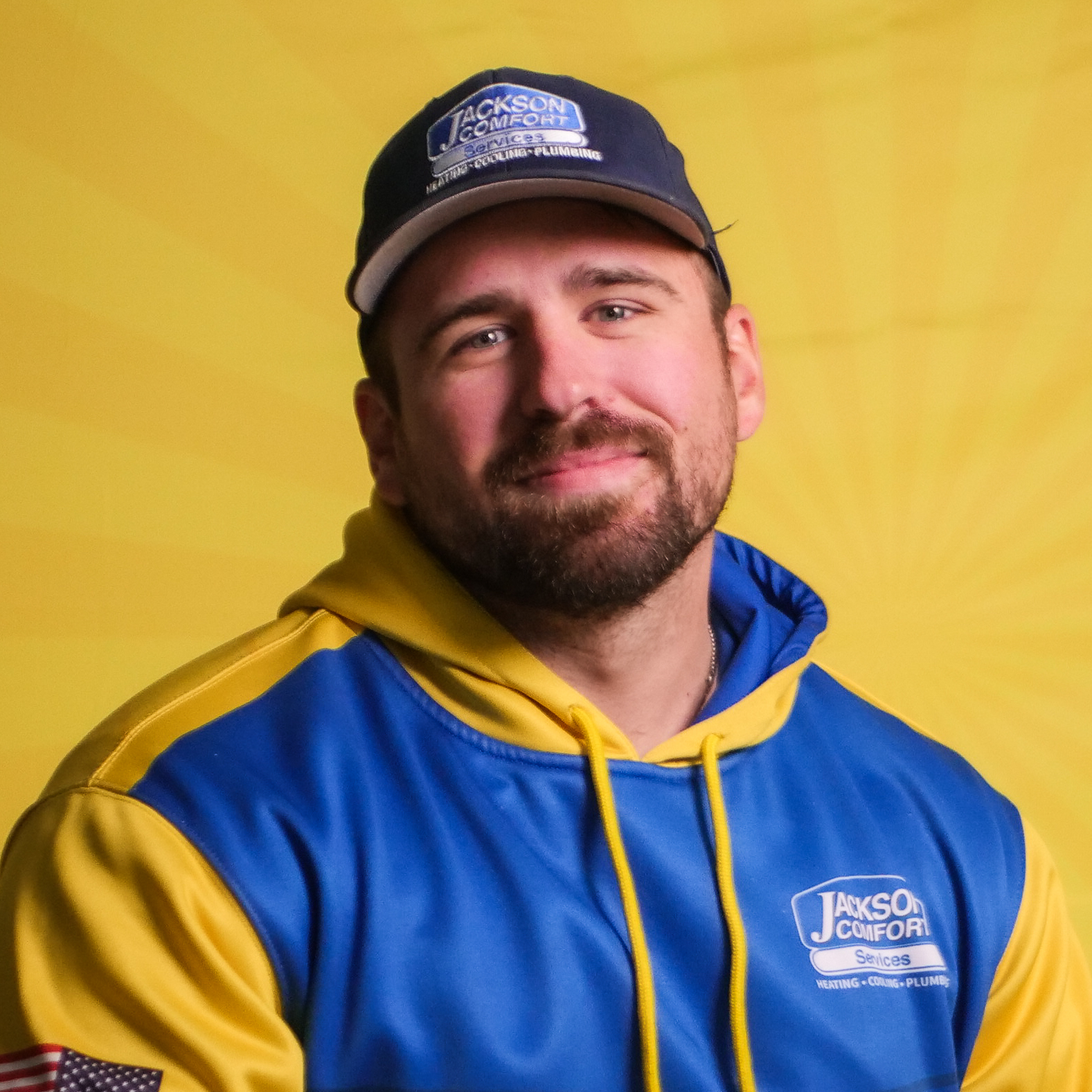 A Member Of Our Team, Dressed In A Blue And Yellow Jackson Comfort Services Uniform And Cap, Poses Confidently In Front Of A Yellow Background.