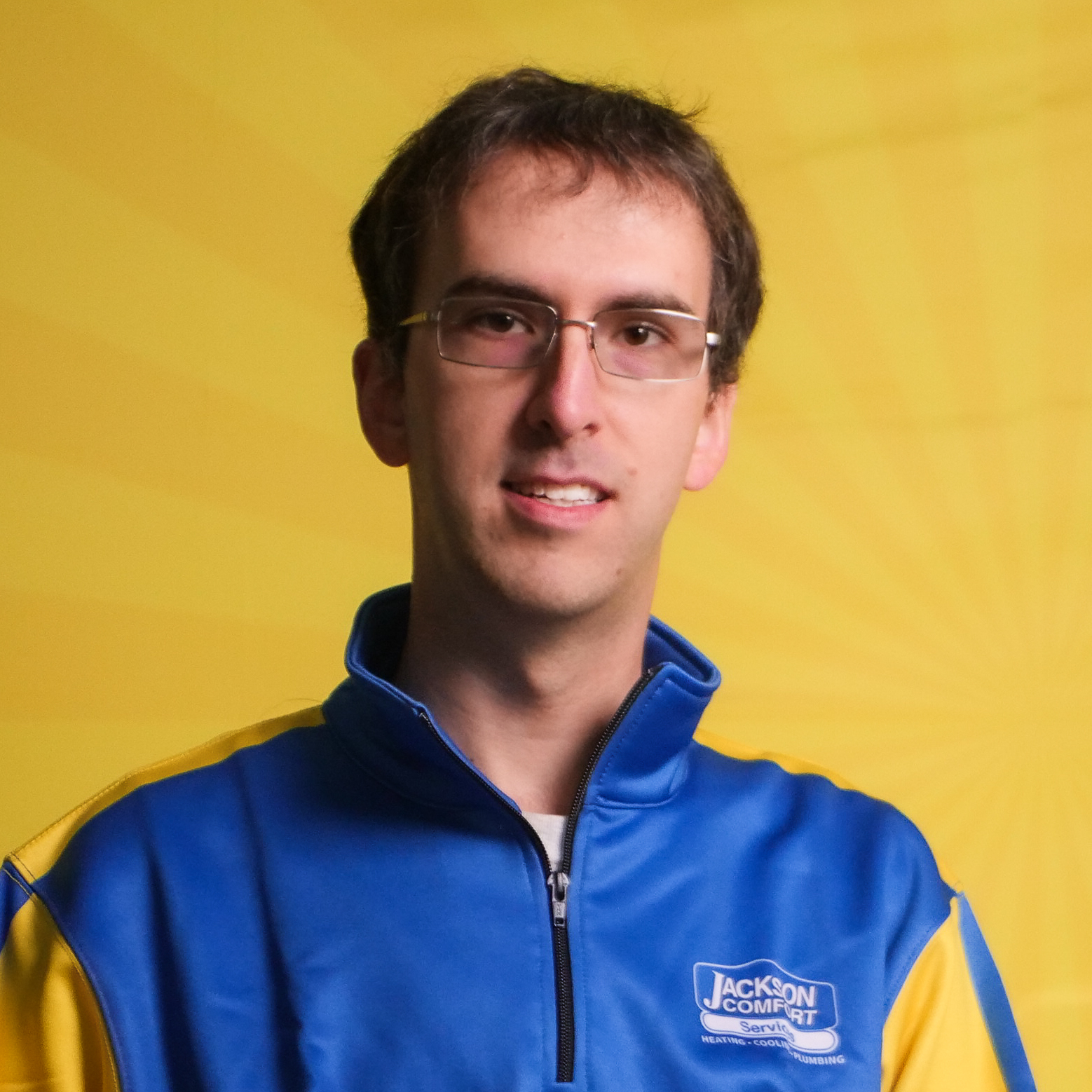 A Member Of Our Team, Wearing Glasses And A Blue And Yellow &Quot;Jackson Comfort&Quot; Jacket, Stands In Front Of A Yellow Background With A Radial Pattern.