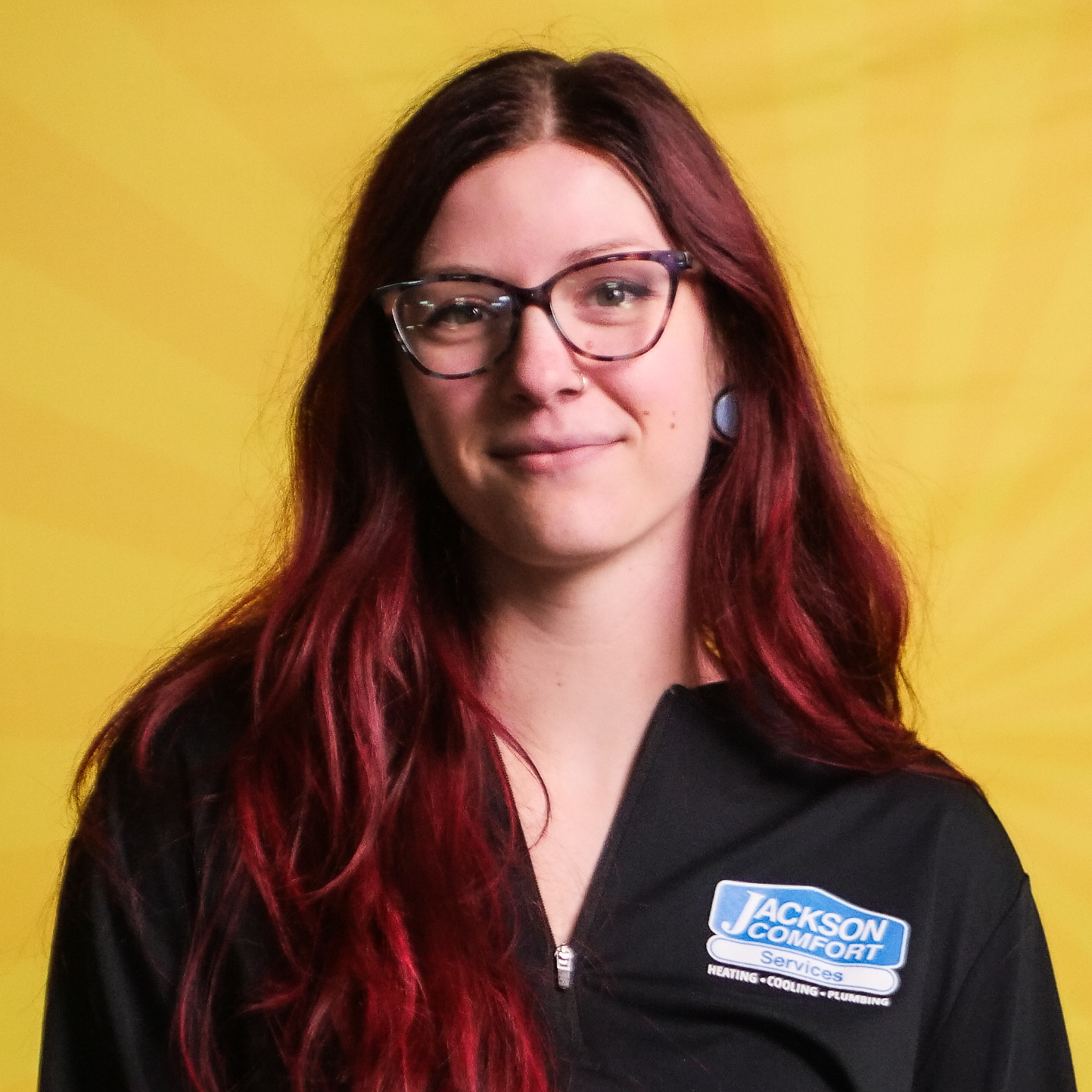 A Member Of Our Team With Long Red Hair And Glasses Wears A Black Jackson Comfort Services Uniform, Standing In Front Of A Yellow Background.