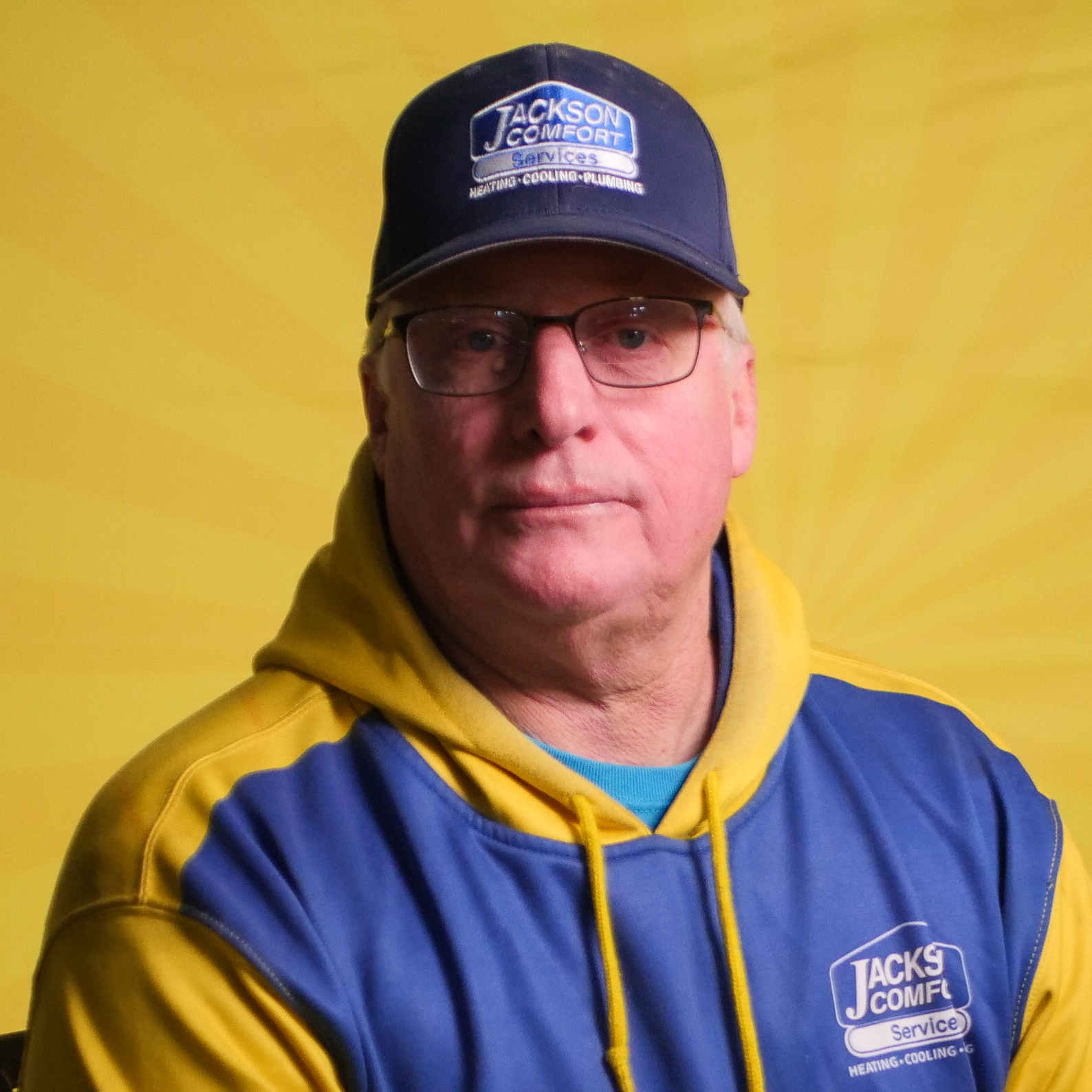 A Member Of Our Team Wears Glasses, A Blue And Yellow &Quot;Jackson Comfort Service&Quot; Hoodie, And A Matching Cap As He Sits In Front Of A Yellow Background.