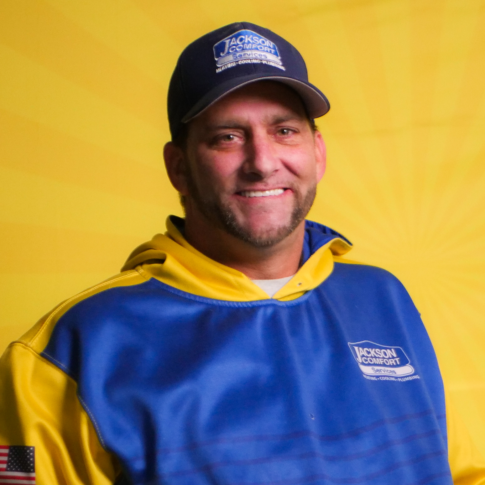 A Member Of Our Team In A Blue And Yellow Jackson Comfort Uniform And Cap Stands In Front Of A Yellow Background, Smiling At The Camera.