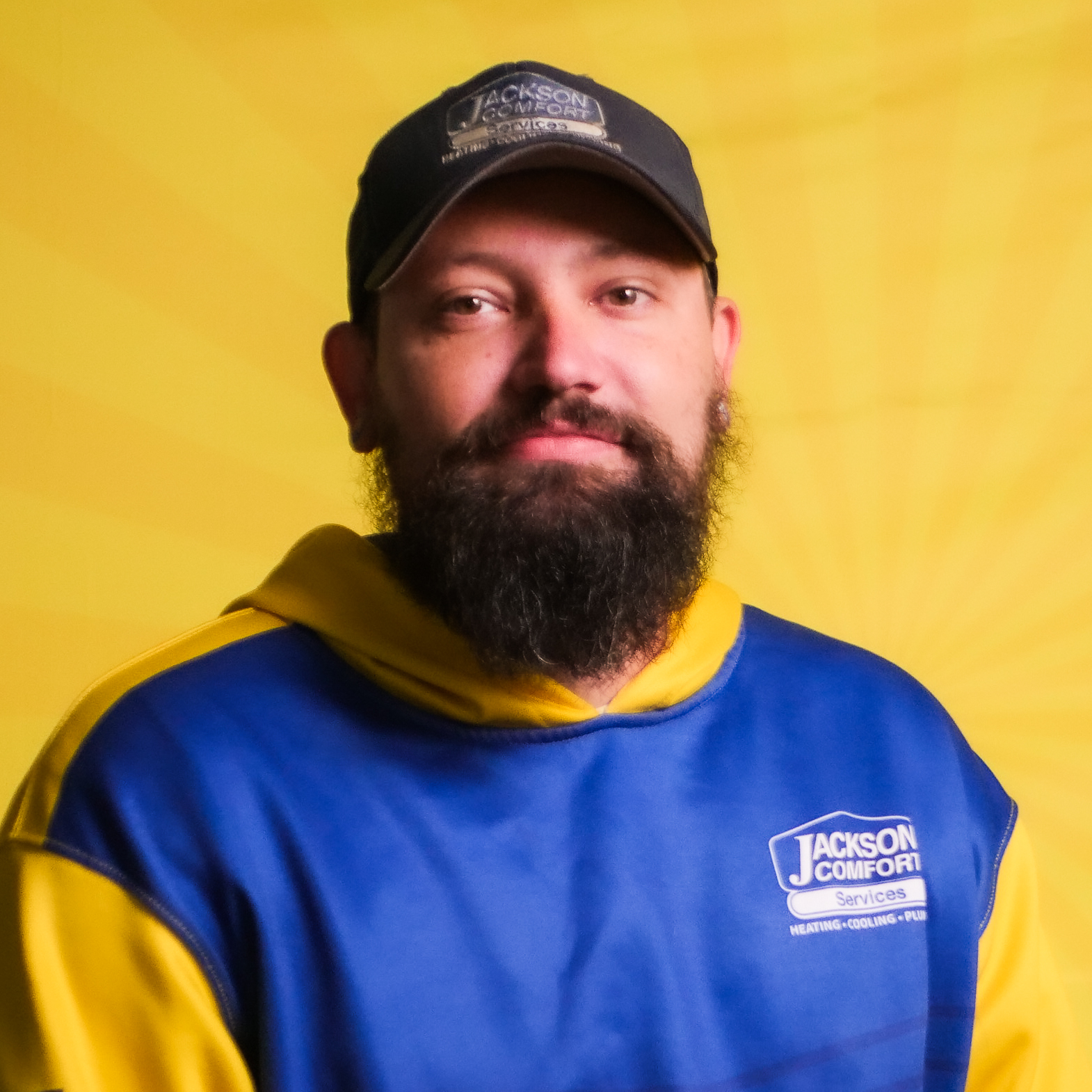 A Bearded Man From Our Team, Wearing A Jackson Comfort Services Hat And Sweatshirt, Poses Against A Yellow Background With A Sunburst Pattern.