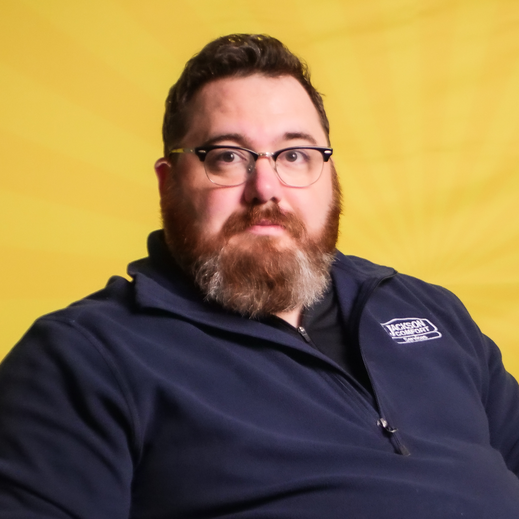 A Bearded Man Wearing Glasses And A Navy Blue Zip-Up Jacket, Representing Our Team, Sits In Front Of A Yellow Background.