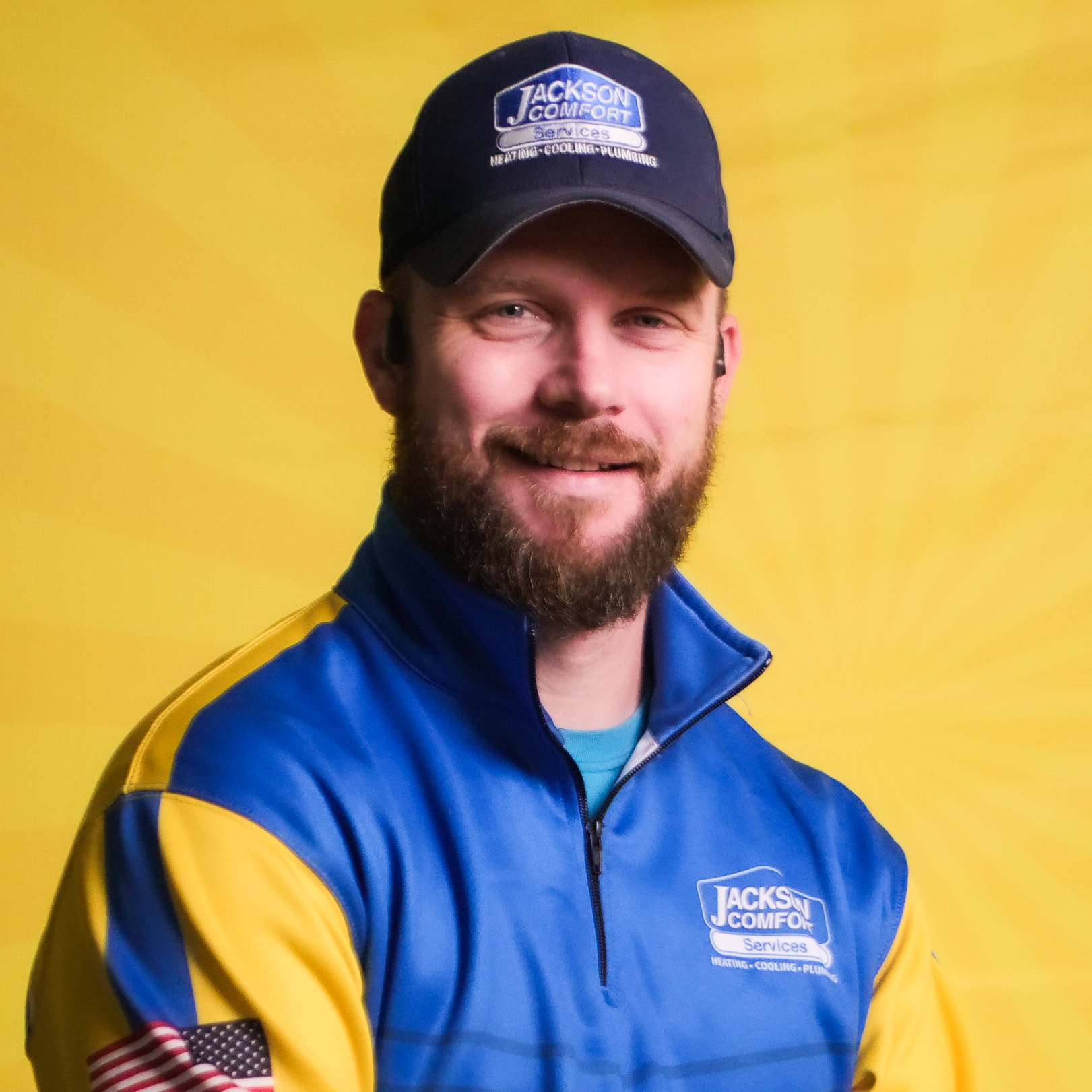 A Bearded Man From Our Team, Dressed In A Blue And Yellow Jackson Comfort Services Uniform And Cap, Stands Confidently In Front Of A Yellow Background.